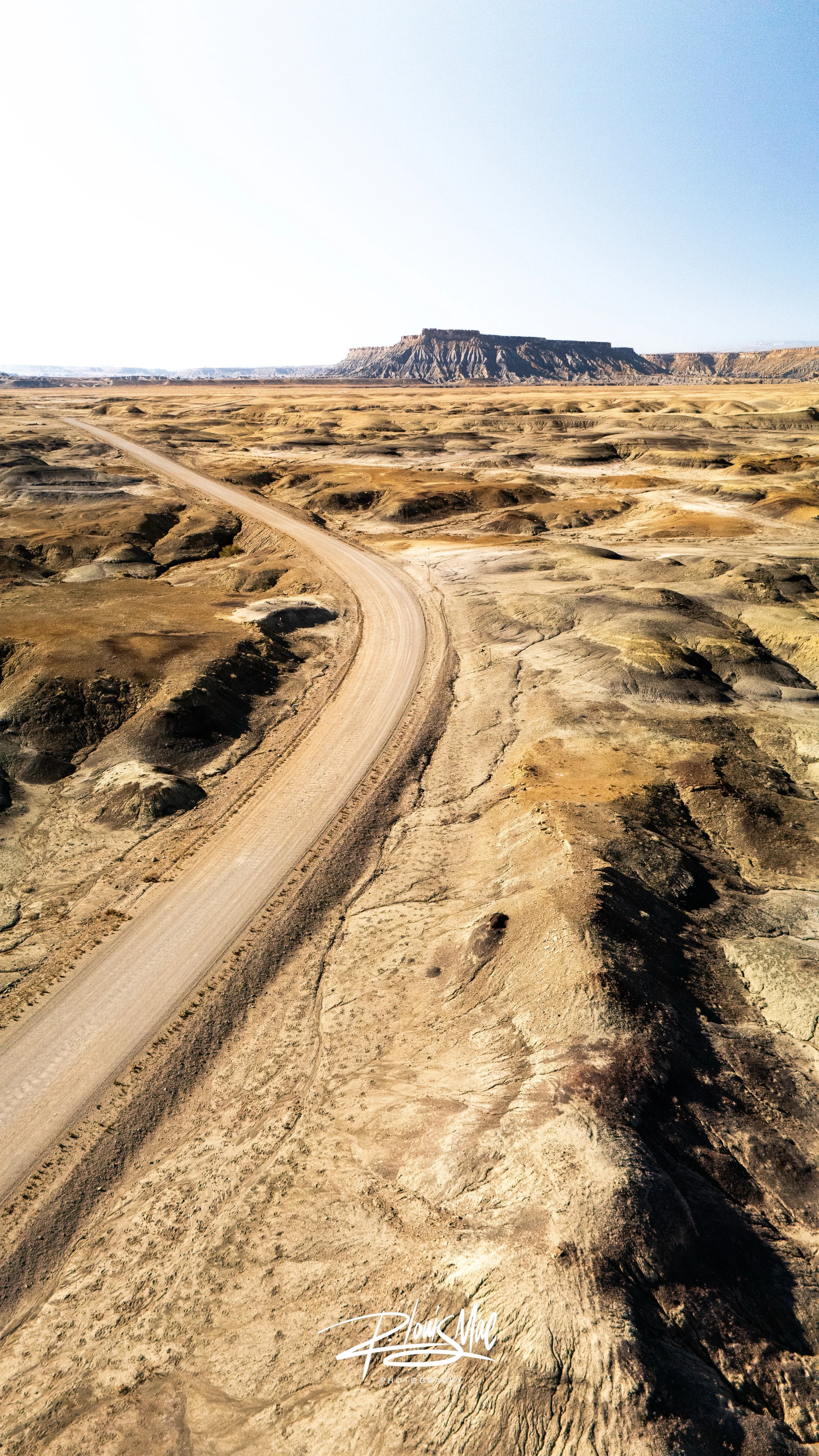 A desert landscape with a winding dirt road and a flat-topped mesa in the distance under a clear blue sky.
