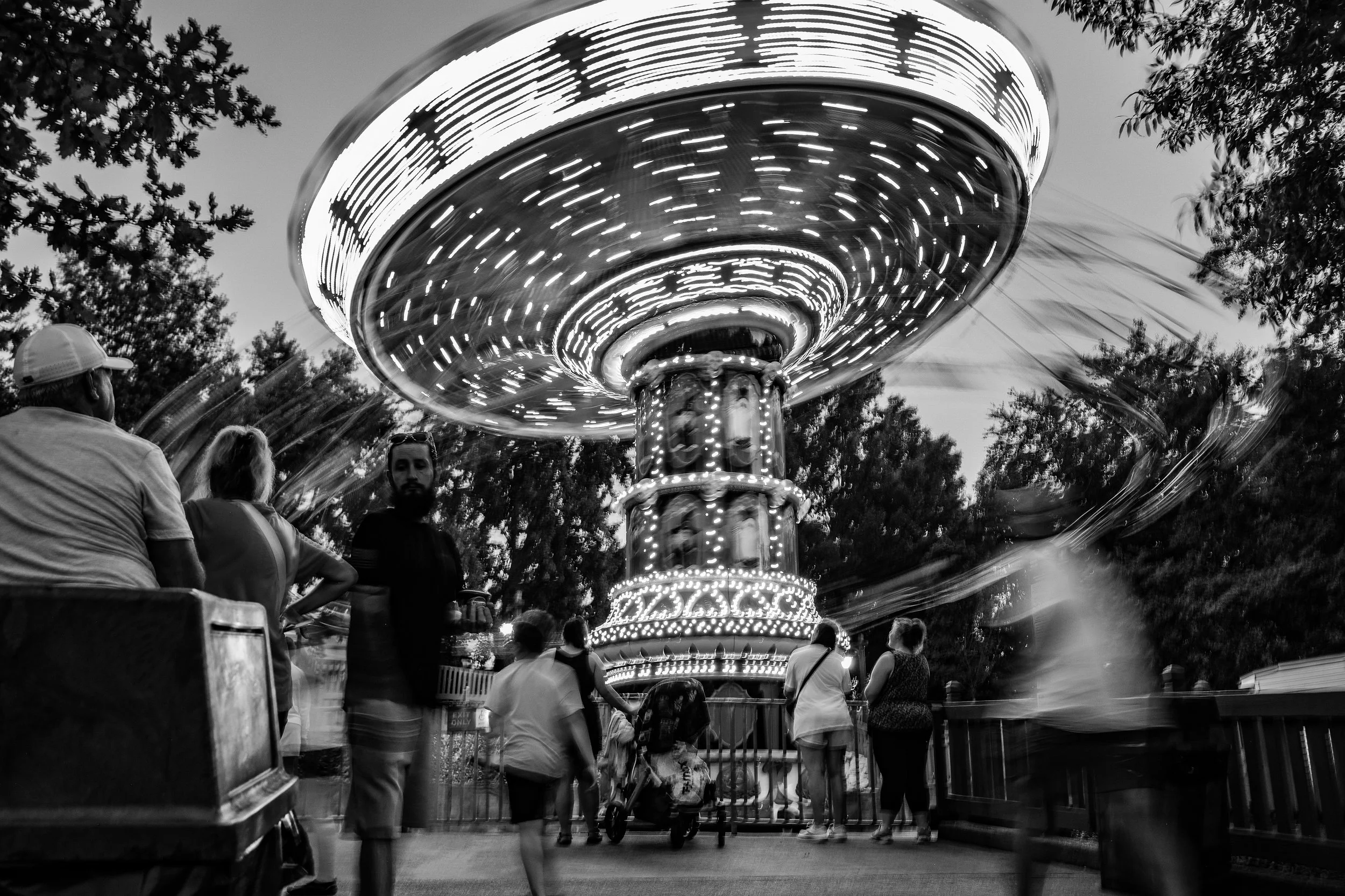 A black and white photo of a carousel ride at a fair, with people standing and walking around. The carousel is spinning with motion blur, and trees are visible in the background.