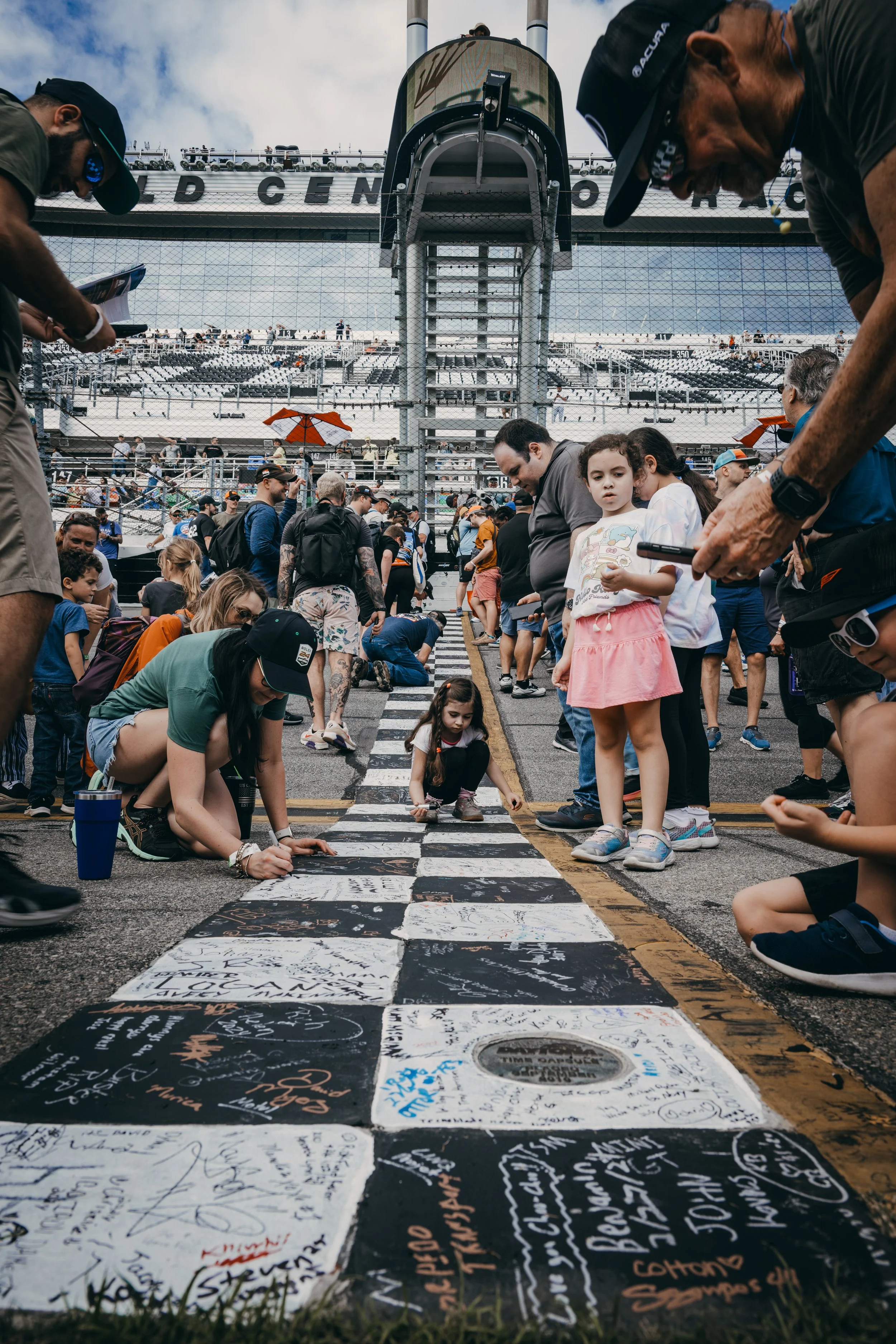People gather on a racetrack, signing and writing messages on large black-and-white checkered tiles in a commemorative event at a racing circuit, with a crowd in the background and a grandstand.
