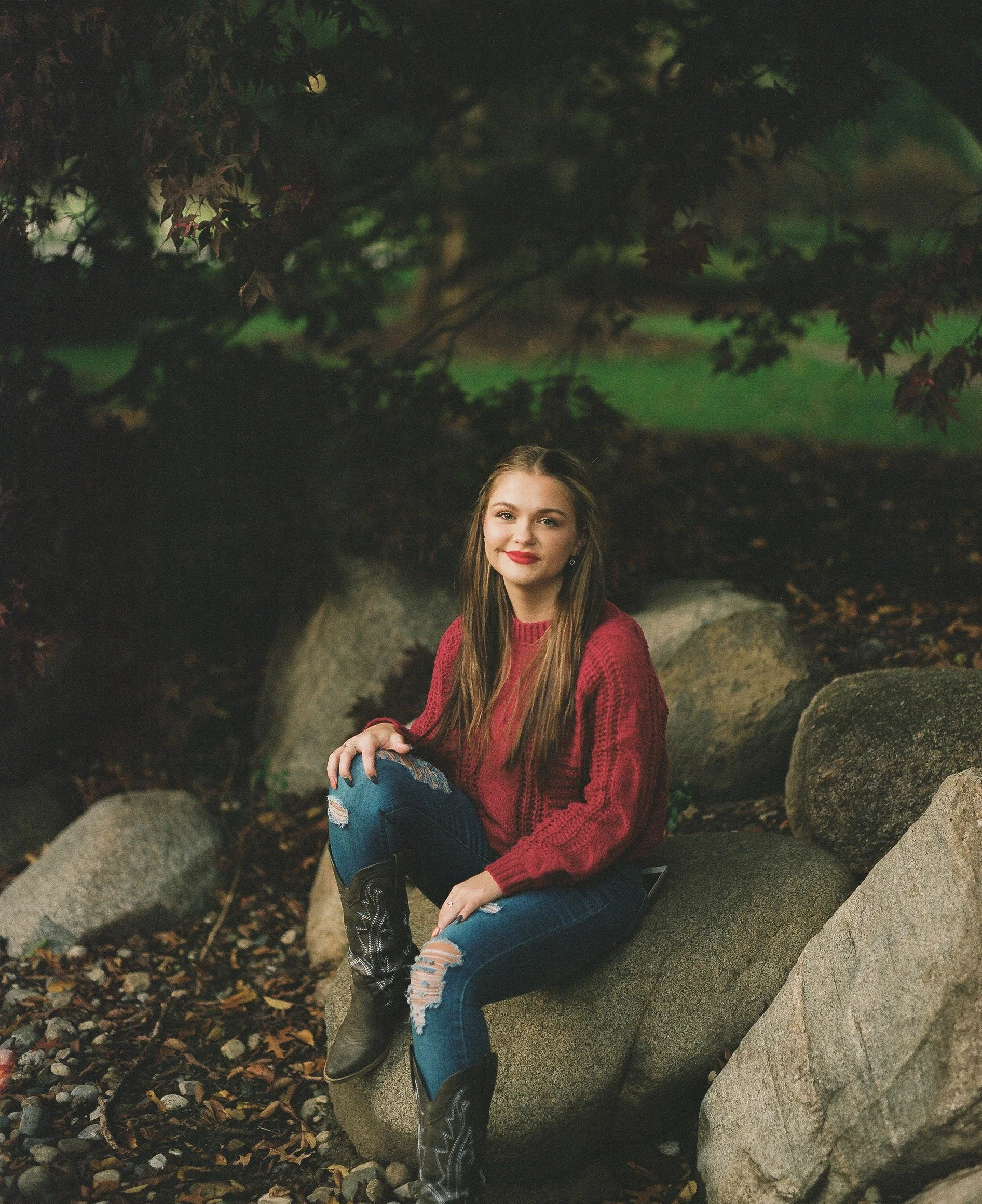 A young woman with long brown hair, wearing a red sweater, ripped blue jeans, and cowboy boots, sitting on a large rock outdoors in a leafy area with trees and fallen leaves.