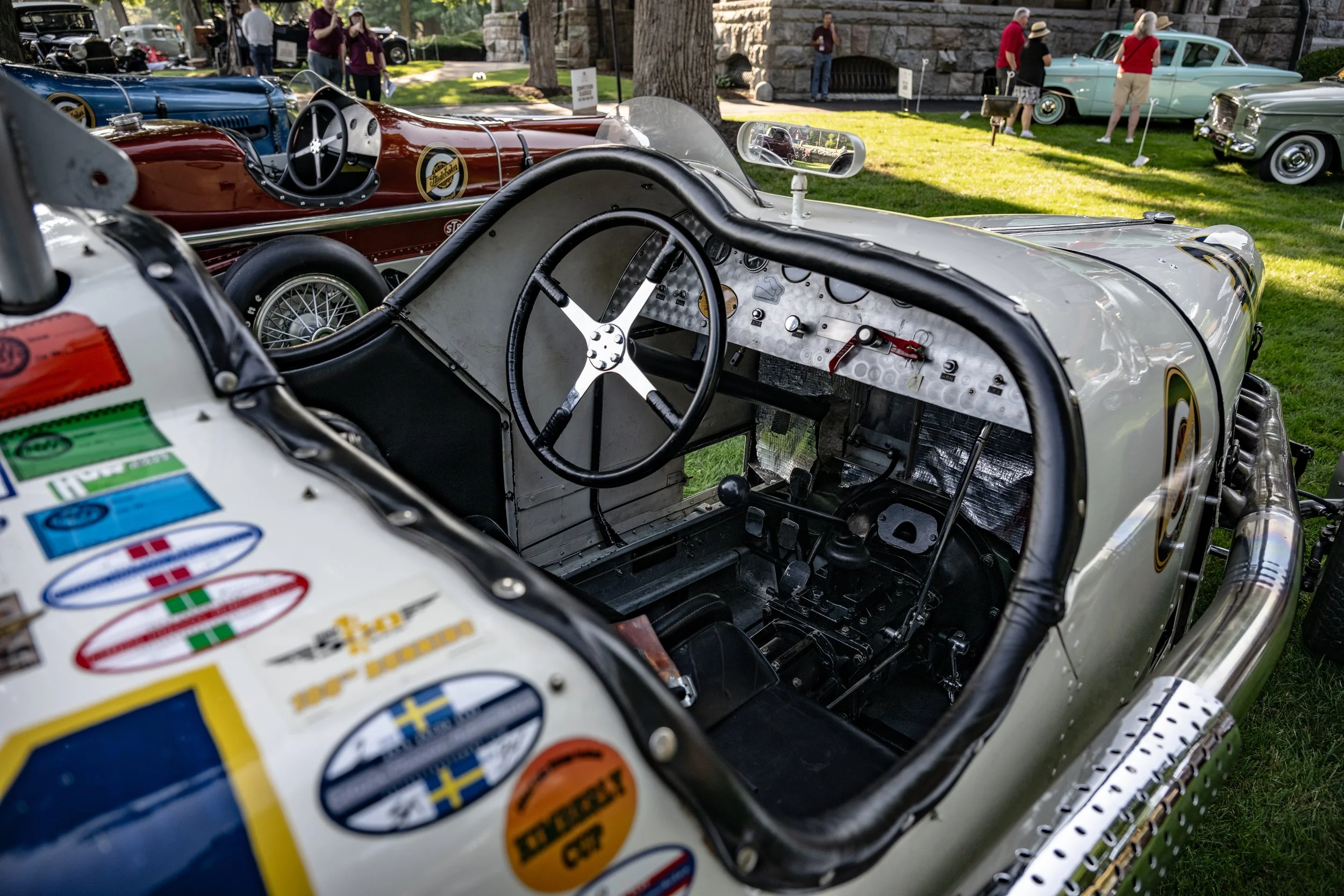 Close-up of a vintage race car interior with a black steering wheel, metal dashboard with gauges, and a black seat, at a classic car show with other vintage cars and people in the background.