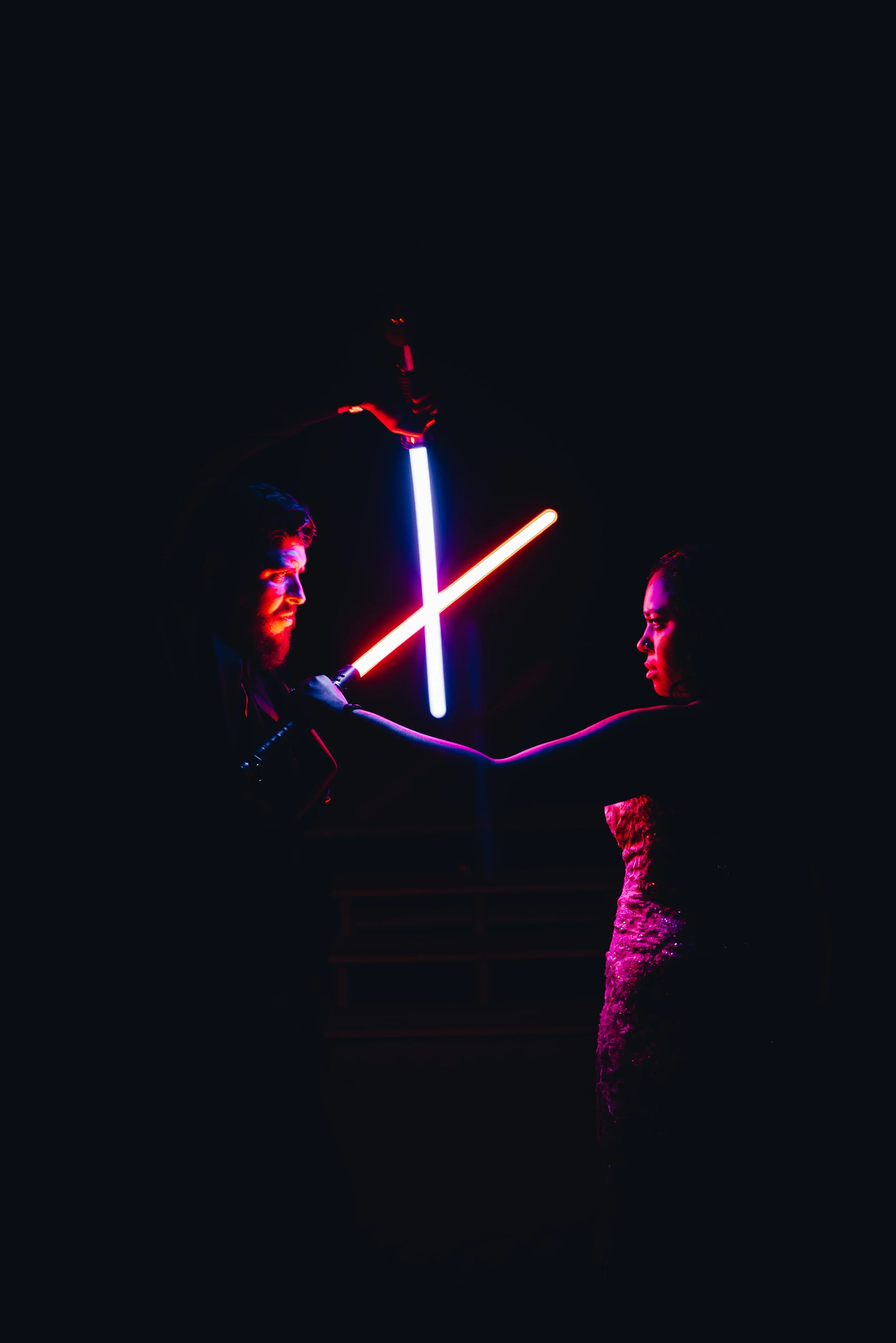 A man with a beard and woman in a dark setting illuminated by neon lights, engaging in a lightsaber duel with glowing pink and blue sabers. The background is mostly dark with minimal detail.