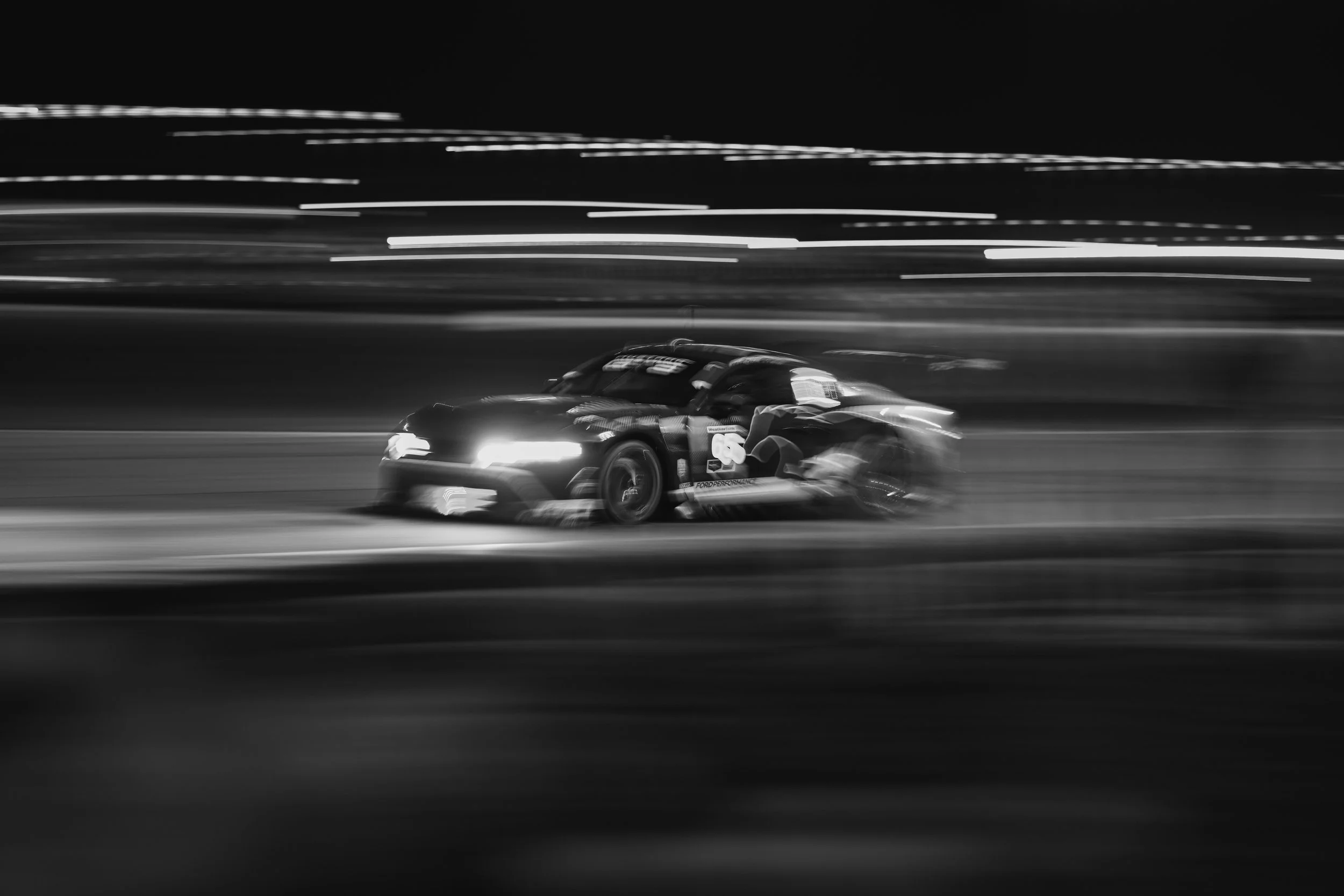 A black and white photo of a race car speeding on a track at night with light trails in the background.