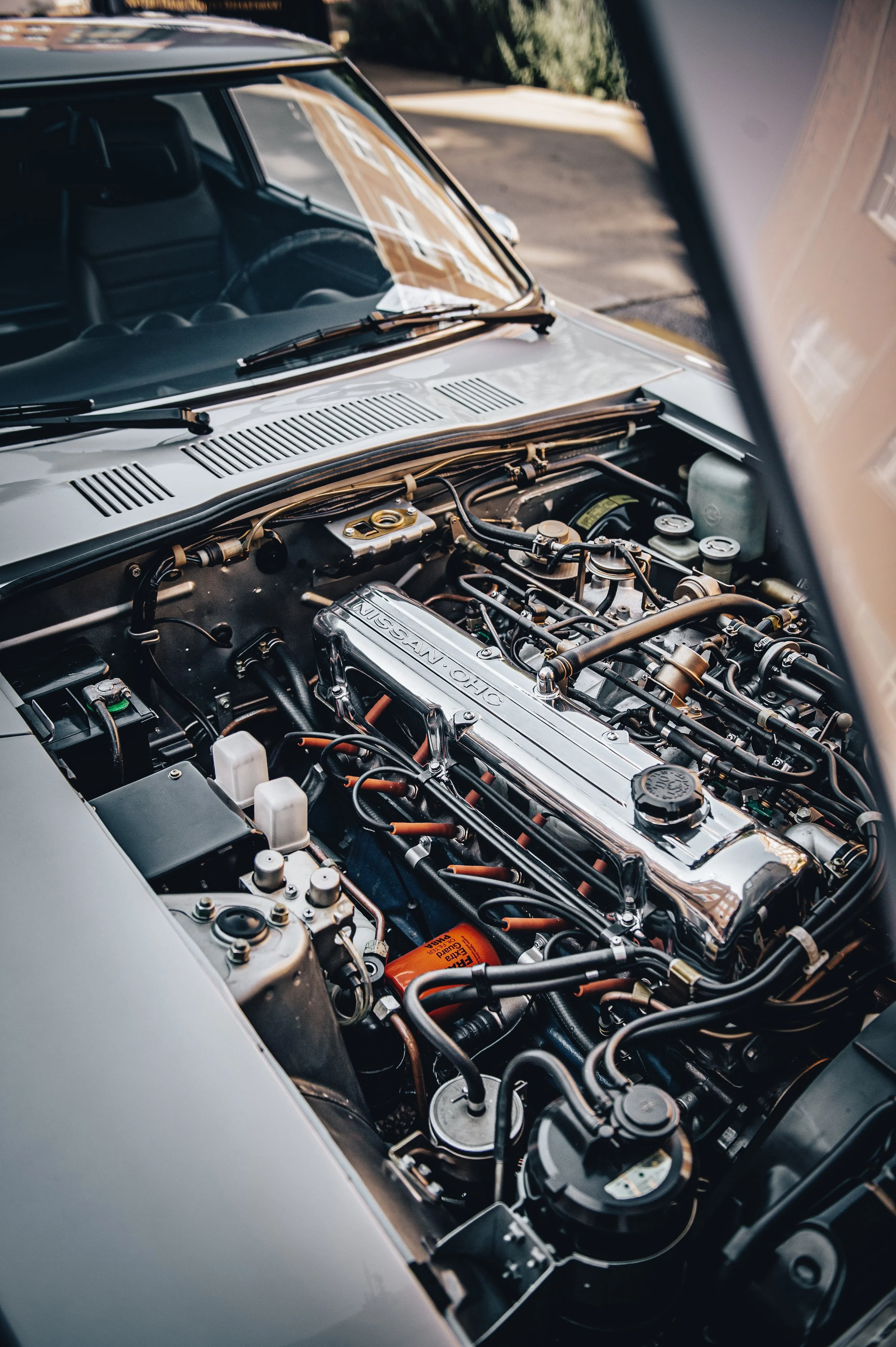 Close-up view of a car engine with a chrome cover labeled 'NISSAN OHC', visible wires, hoses, and engine components.