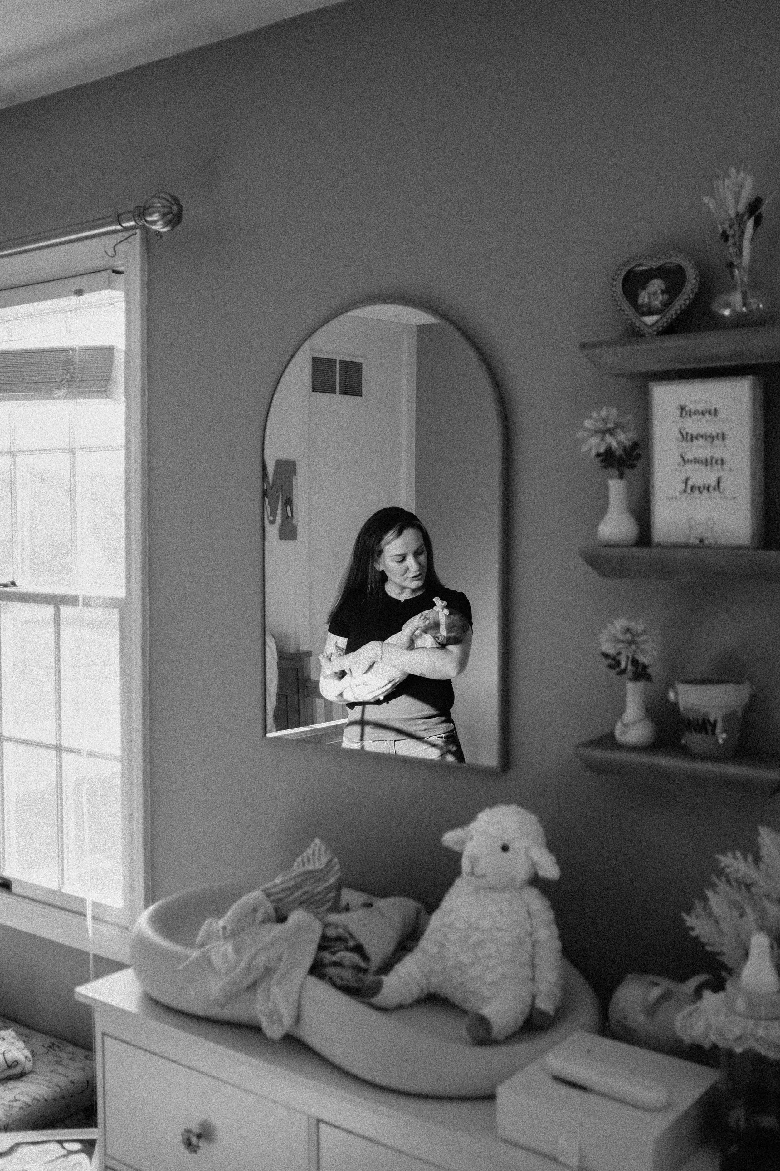 A woman holding a newborn baby, seen through a mirror in a nursery decorated with toys, framed pictures, and flowers.