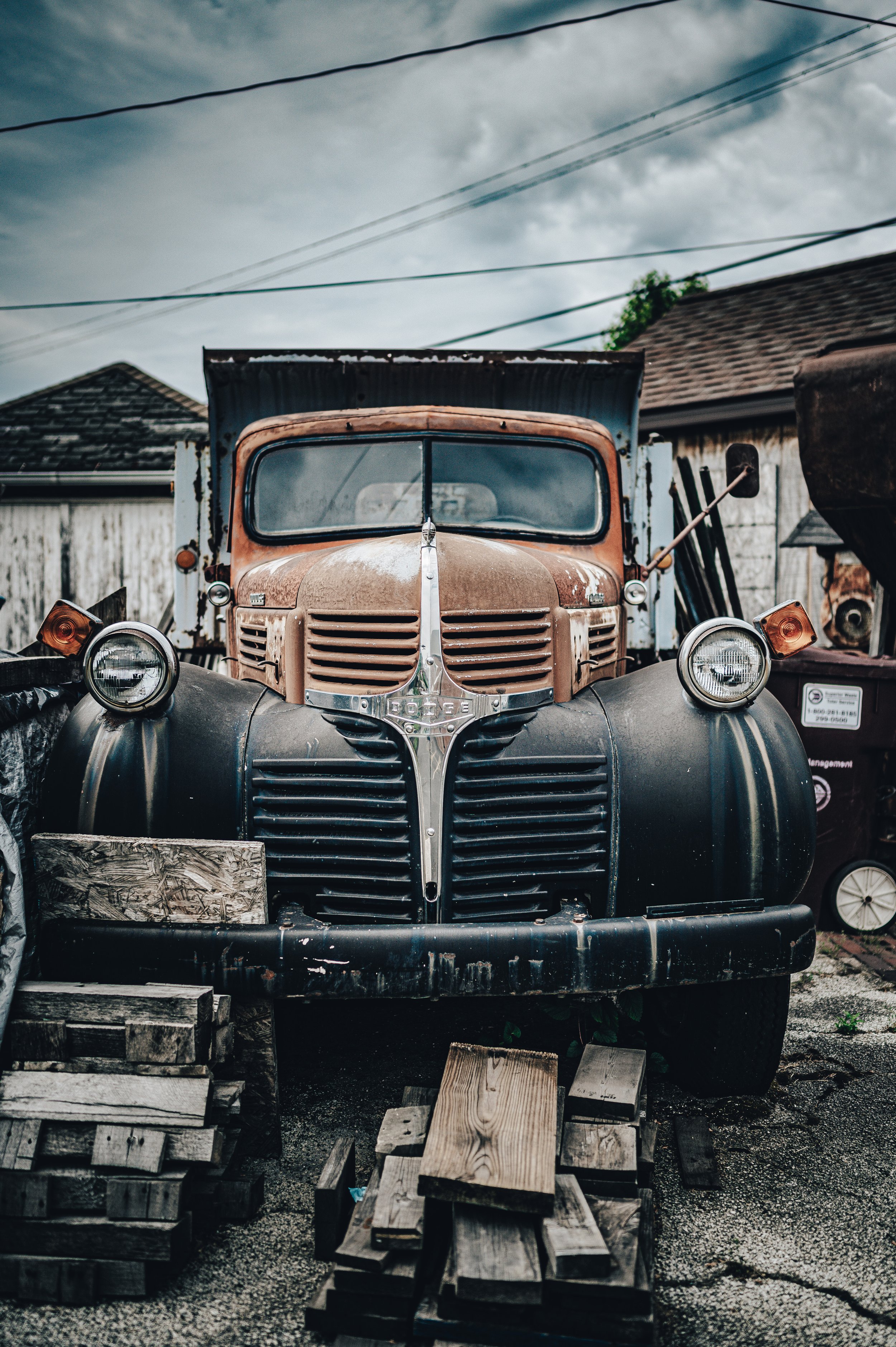 Old rusty vintage Dodge truck with black fenders parked outdoors on a gravel ground, surrounded by wooden pallets, under cloudy sky.
