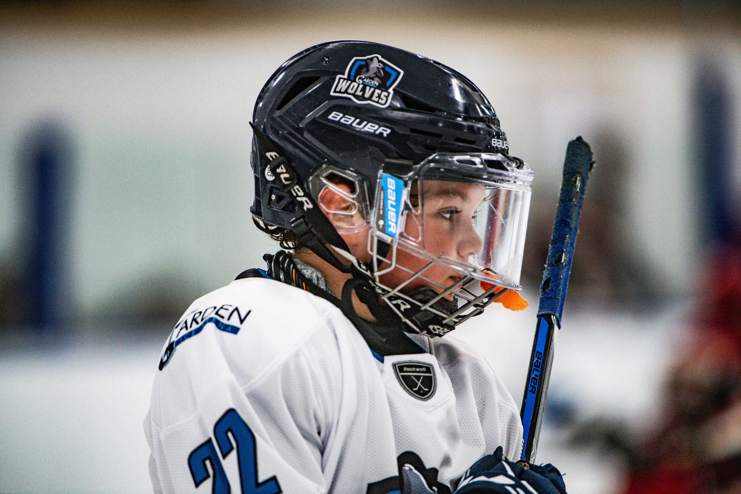 Close-up of a young hockey player wearing a black helmet with a face shield, a white jersey with the number 22, and holding a blue hockey stick on an ice rink.