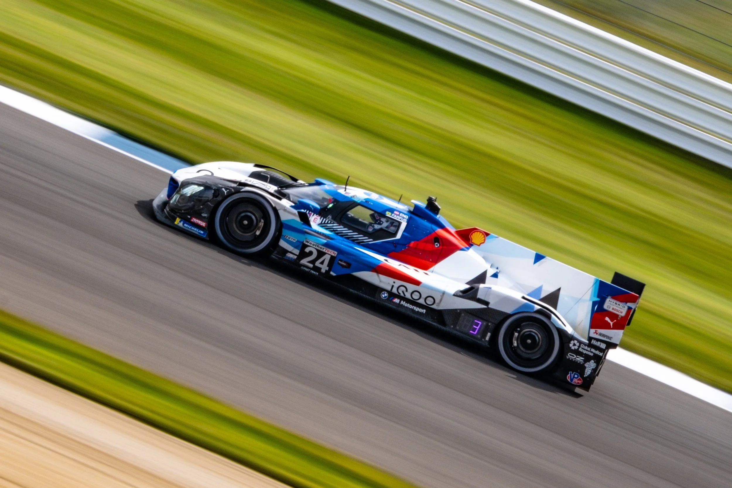 A race car moving at high speed on a racetrack with a blurred background, featuring racing sponsors and a blue, white, red, and black color scheme.