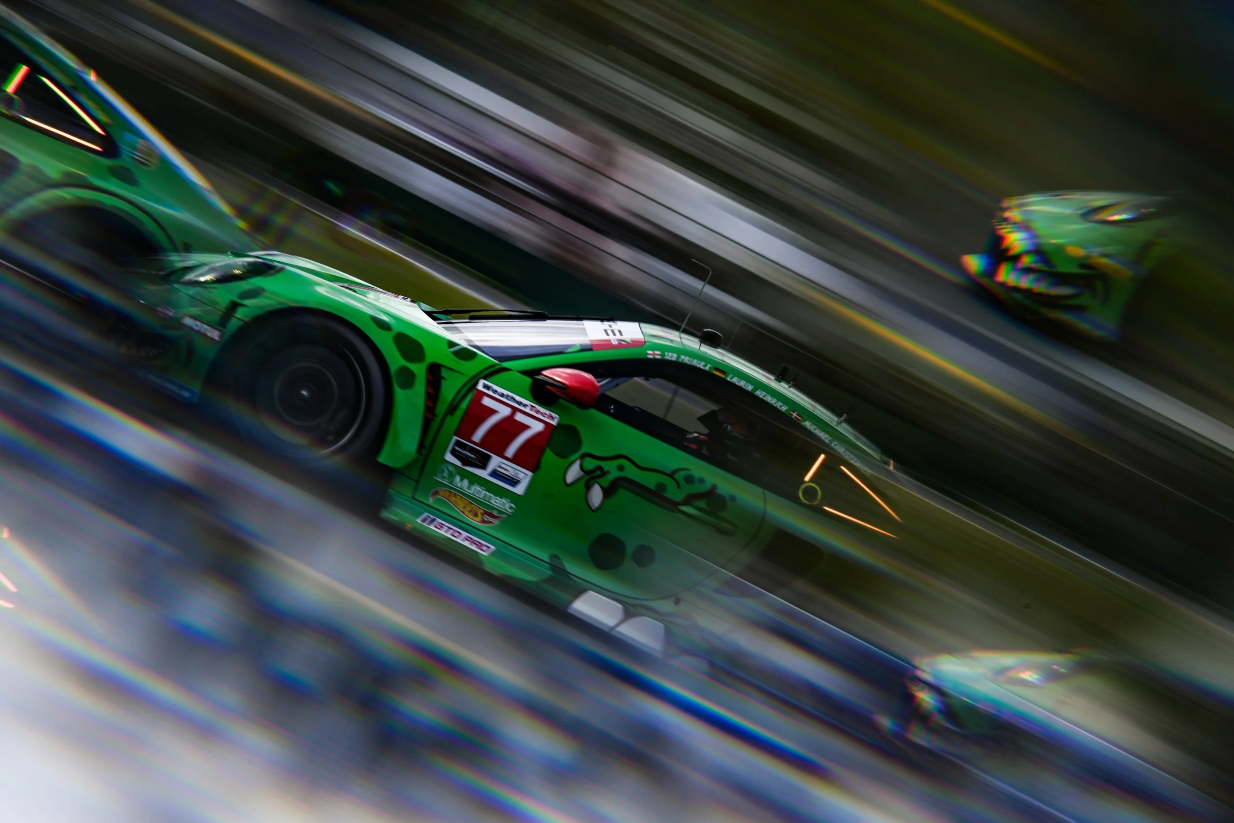 Green racing car with the number 77 and sponsor logos, racing on a track with motion blur effect.