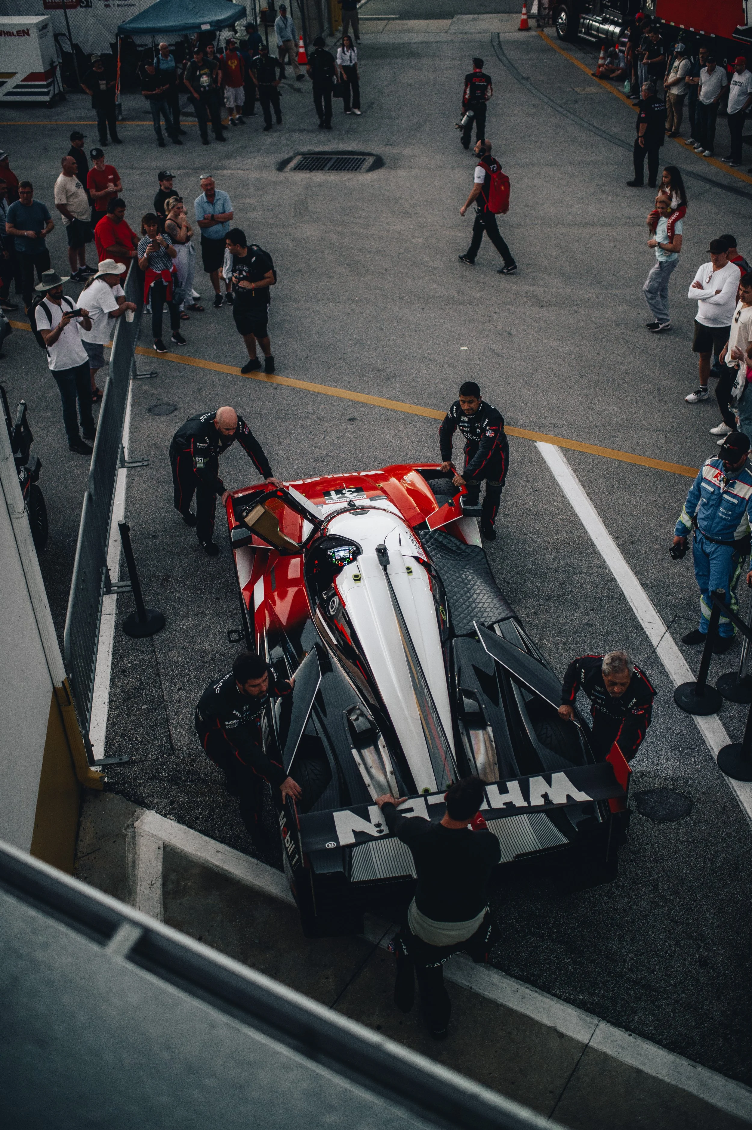 Race car pit stop with crew members working on a sleek, aerodynamic racing vehicle surrounded by spectators at a race track.