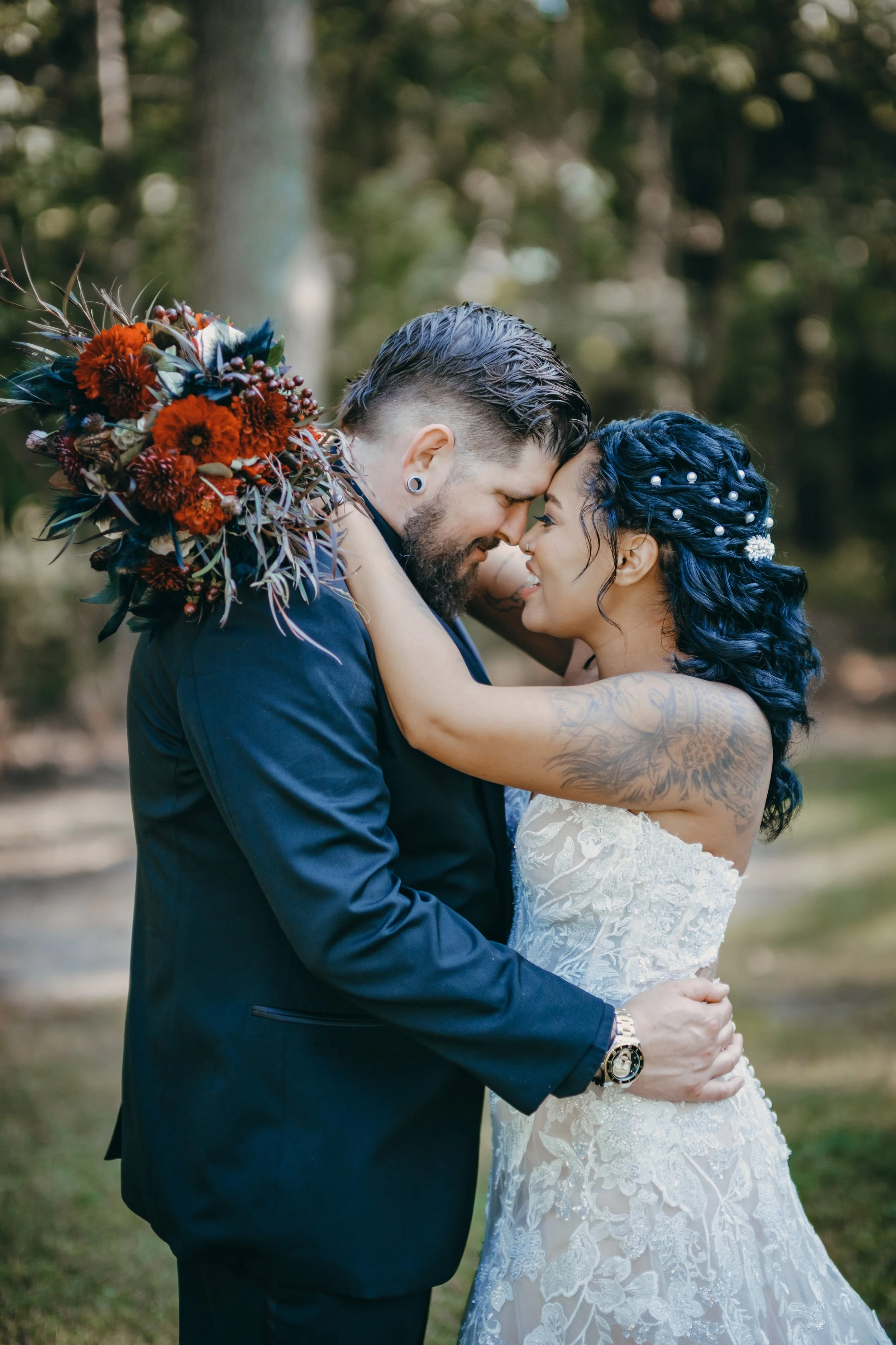 A couple on their wedding day embracing outdoors, with the groom in a navy suit and the bride in a white lace wedding dress, holding a bouquet of red flowers.