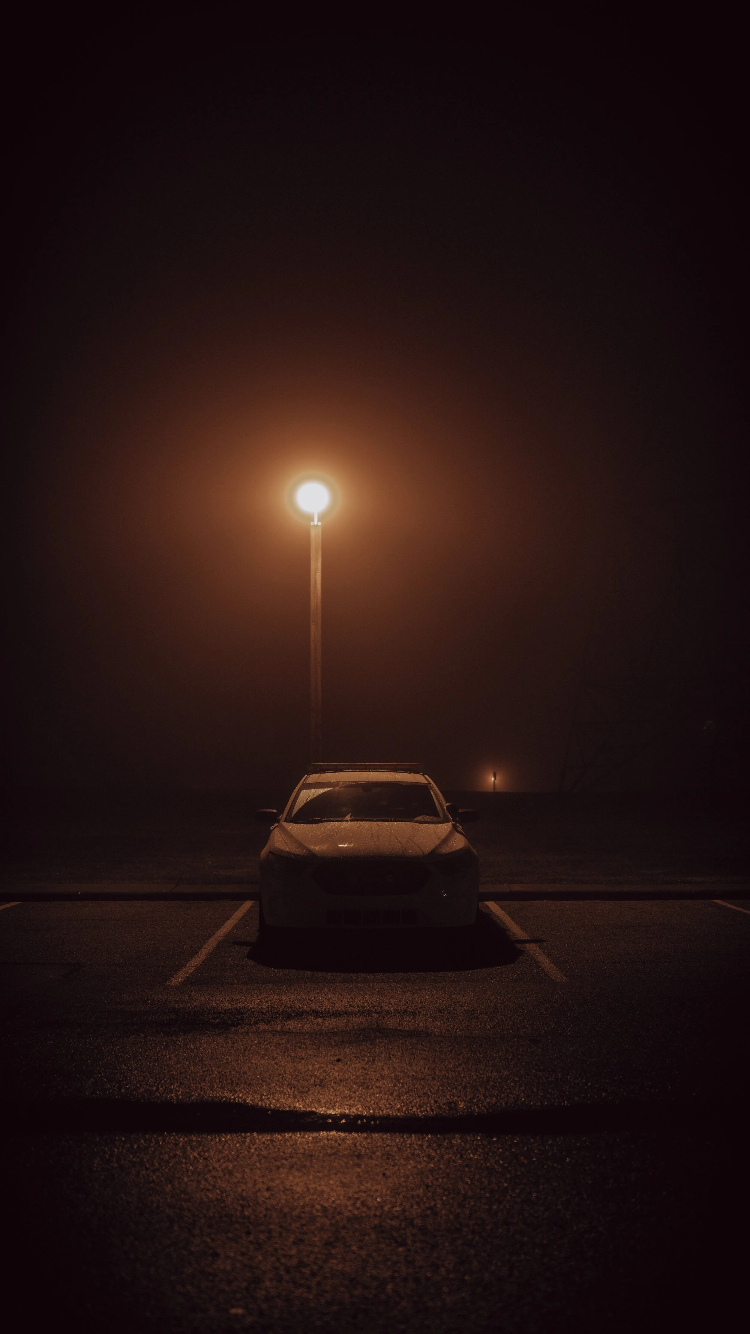 A car parked at night in an empty parking lot with a bright streetlamp illuminating the foggy night, creating a glowing halo around the light.