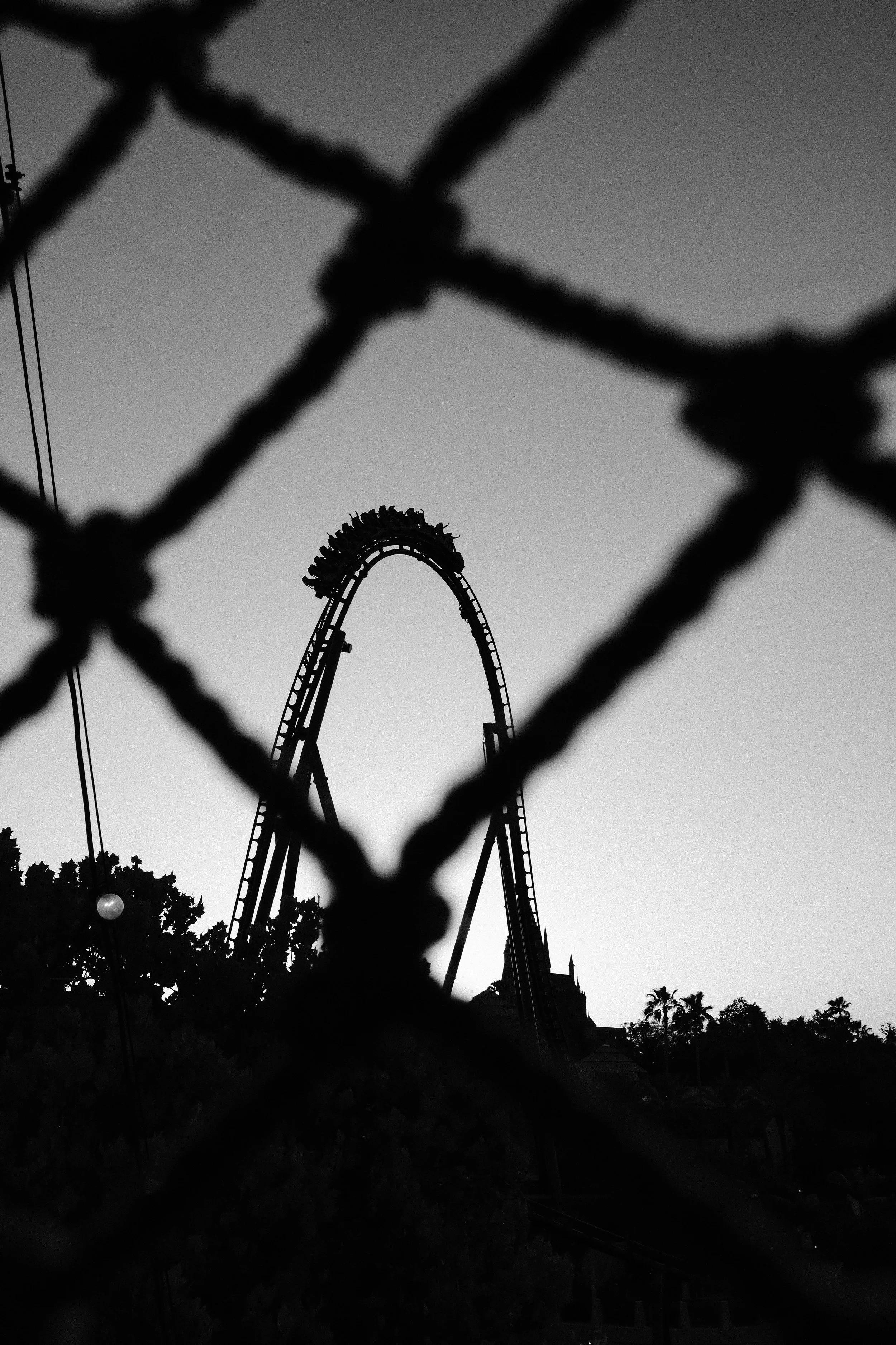 Black and white photo of a roller coaster seen through a chain-link fence at sunset or dusk.
