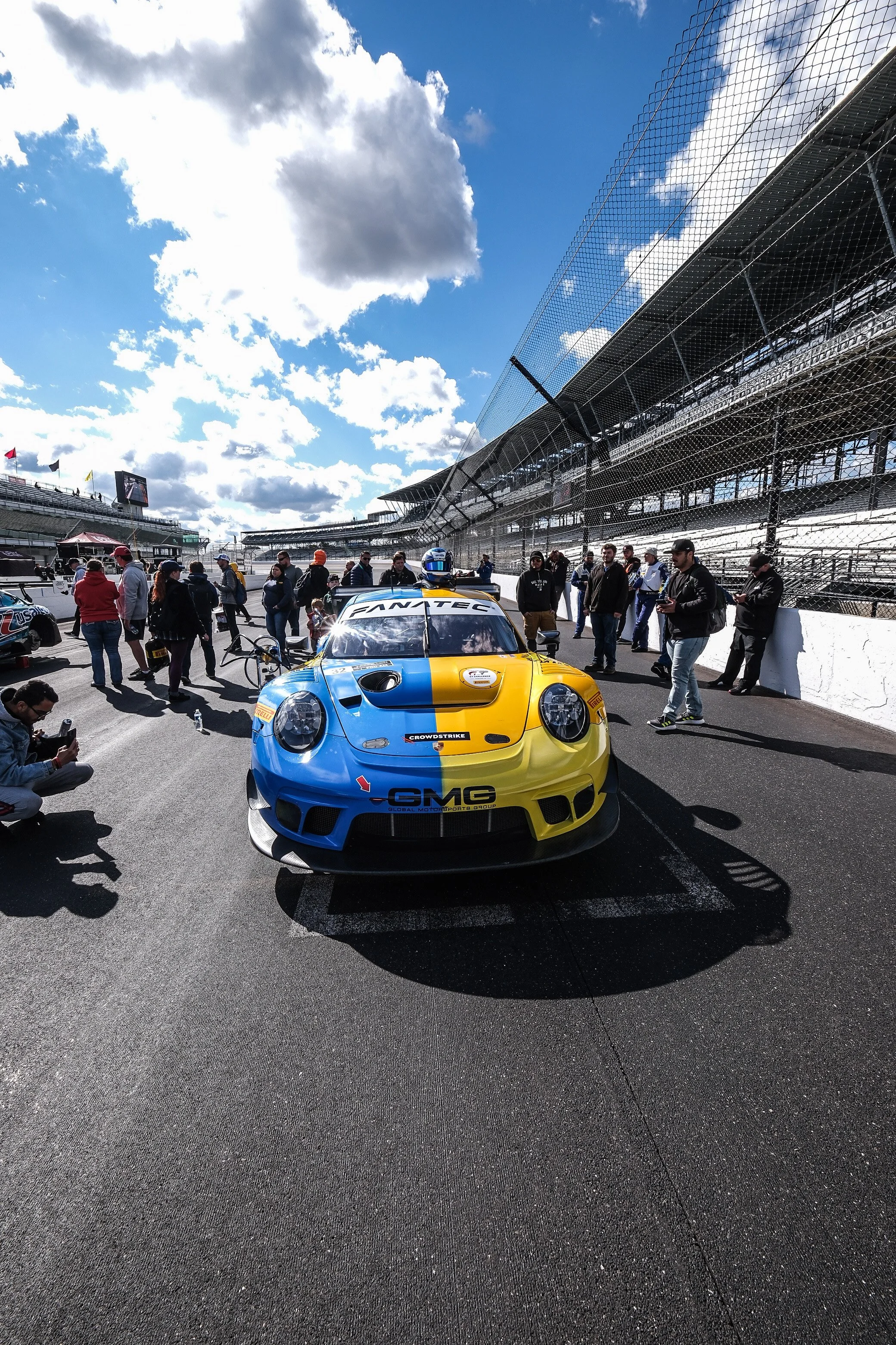 A blue and yellow race car on a race track with people standing around and a large grandstand with a safety fence in the background, under a partly cloudy sky.