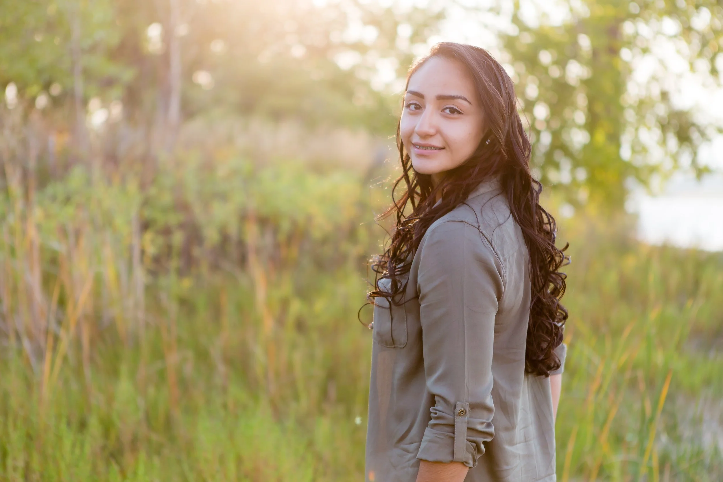 Young woman with long curly hair smiling outdoors in a natural setting with trees and green foliage.