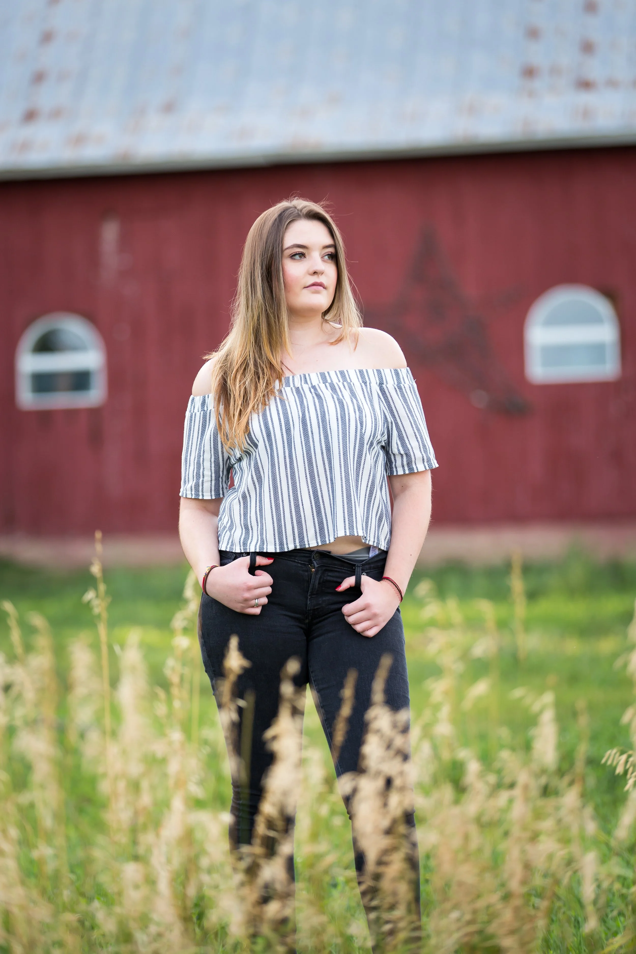An outdoor high school senior portrait session of a young woman standing in a grassy field with a classic red barn in the background on a private farm in Boulder, Colorado.