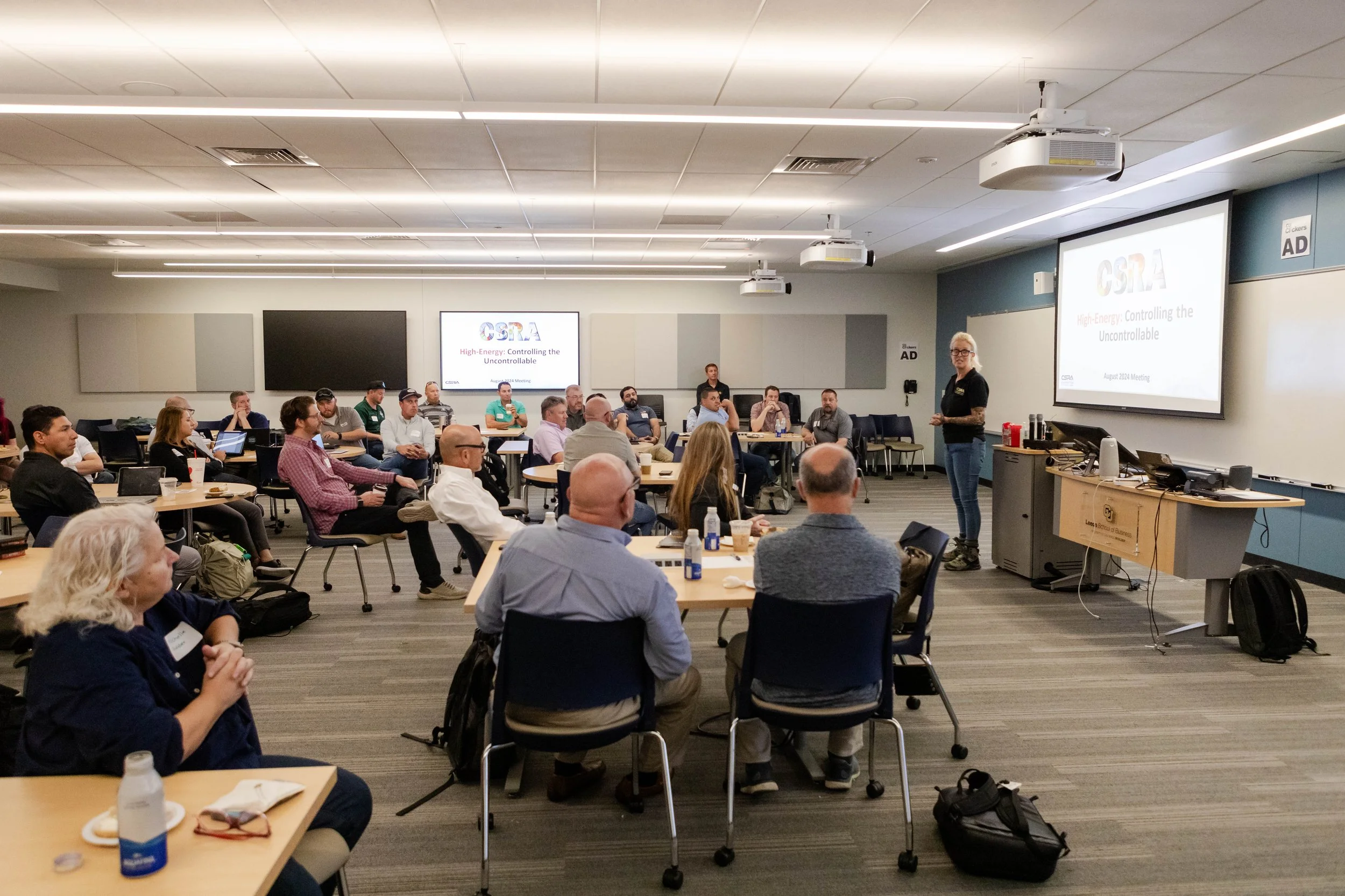A conference room filled with attendees sitting at tables, listening to a speaker at the front. The speaker is a woman with glasses, standing near a large screen displaying a presentation titled 'High Energy: Controlling the Uncontrollable.' The room