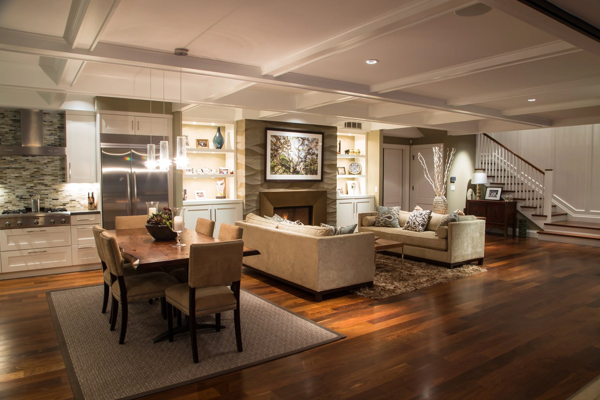 A spacious, modern living room and kitchen area with hardwood floors, a dining table with chairs, beige sofas, a fireplace, and built-in shelving with decor and photos. Large ceiling with recessed lighting.