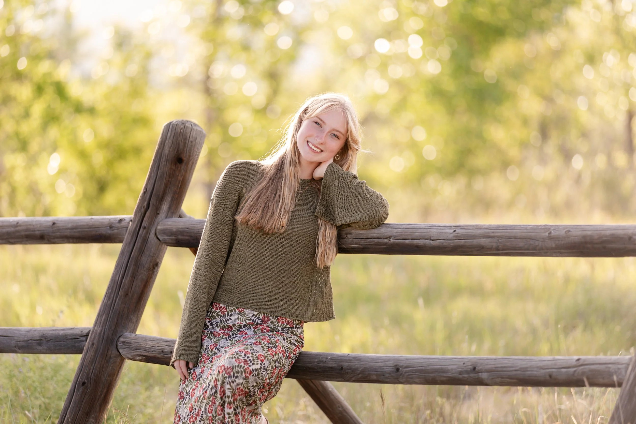Golden hour senior picture at Coot Lake featuring a smiling high school senior in a green sweater leaning against a rustic wood fence with warm evening backlighting.