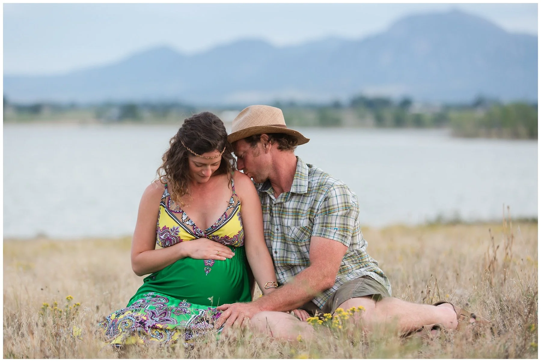 A pregnant woman and a man sitting together outdoors near a lake with mountains in the background. The woman is touching her belly, and the man is leaning in close with his head touching hers.