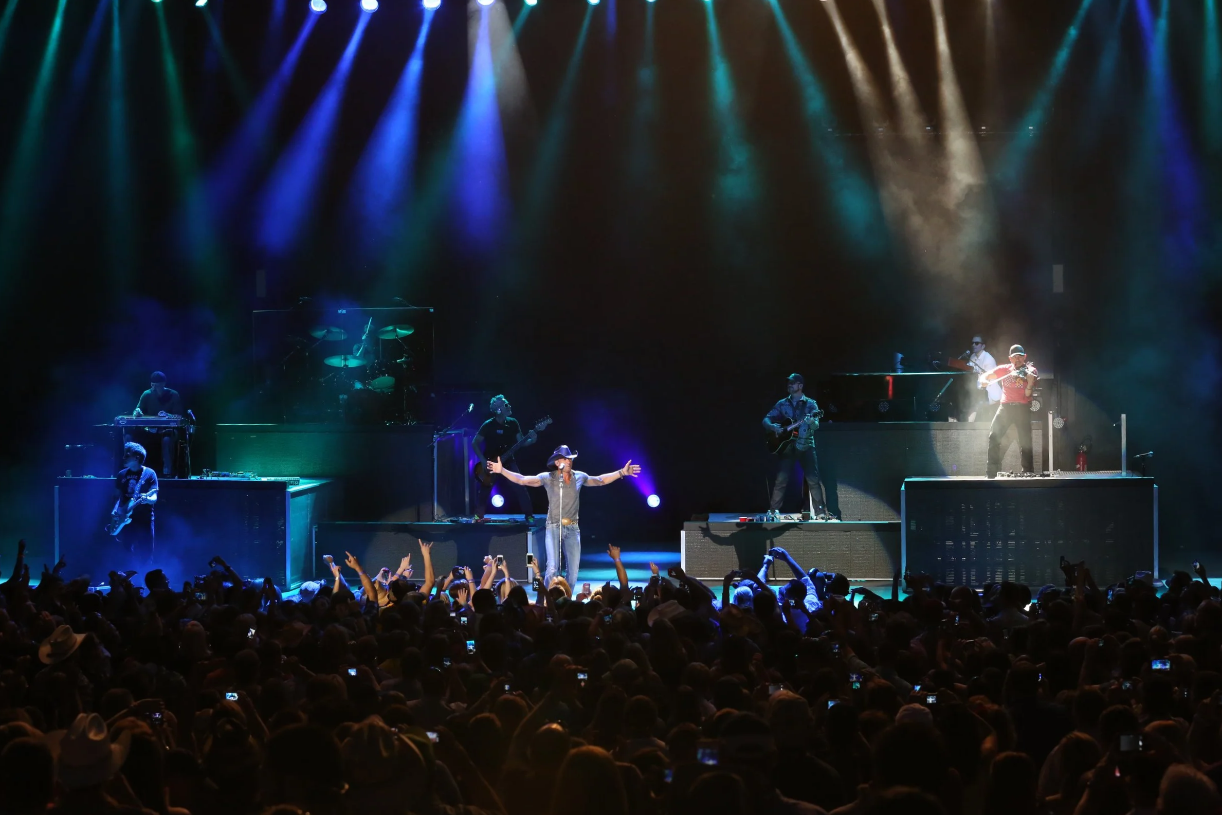 A live concert on a large stage with multiple musicians performing. There are colored lights shining down, and the audience is visible in the foreground with many raising their hands, cheering, and some taking photos or videos.