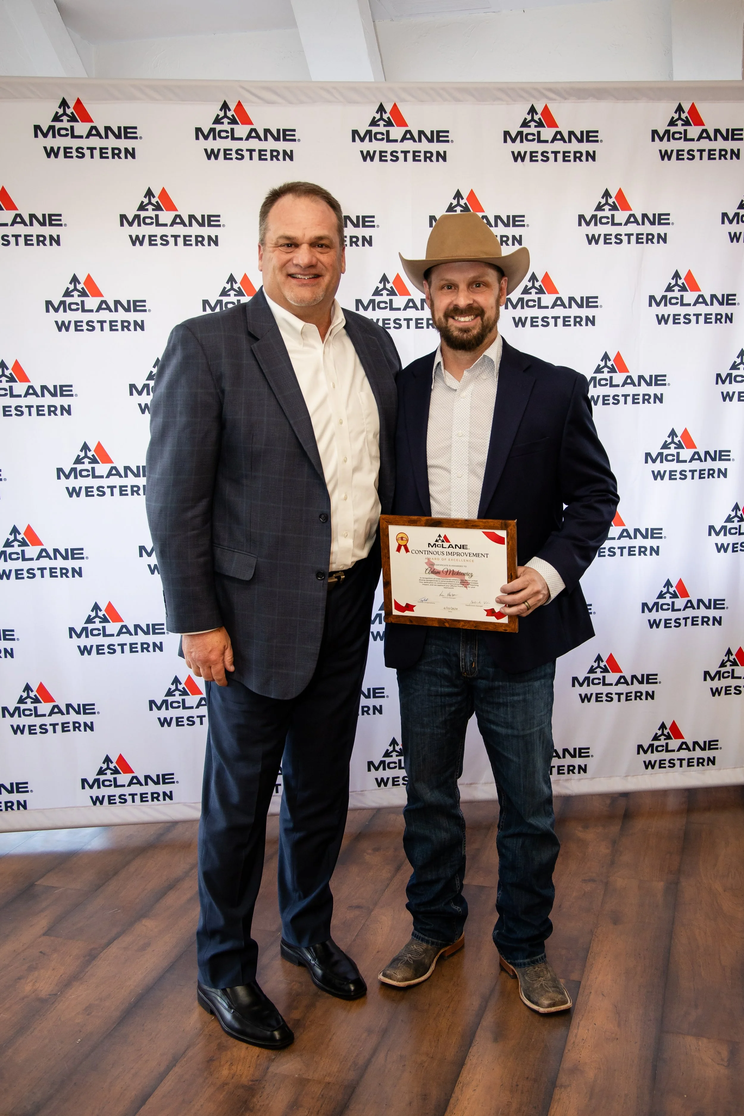 Two men standing together in front of a McLane Western branded backdrop, one is holding a framed award. The man on the left is dressed in a plaid suit with a white shirt and black shoes. The man on the right is wearing a cowboy hat, a dark blazer, a 