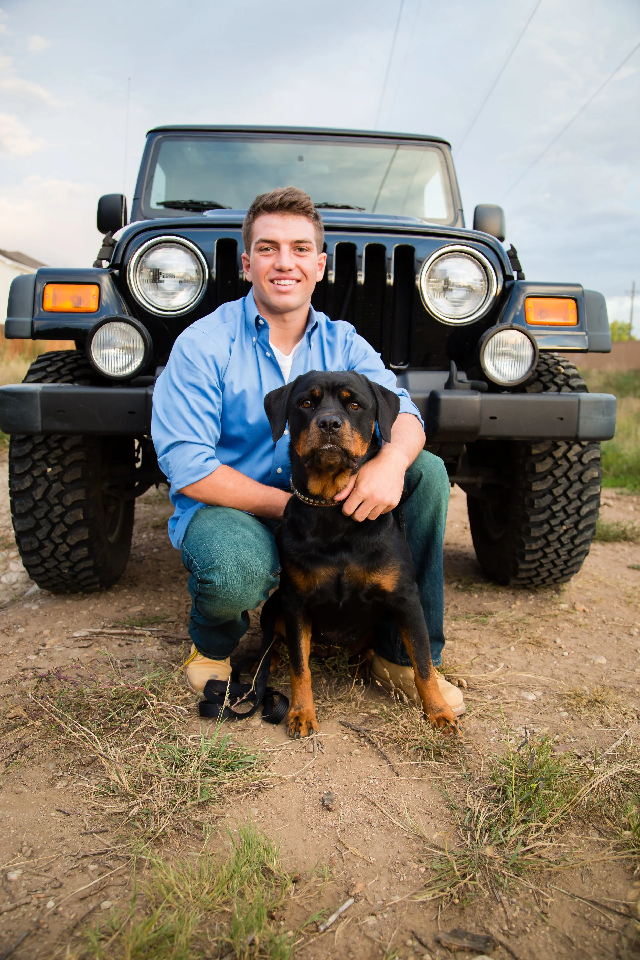 A personalized high school senior lifestyle portrait session featuring a young man crouching with his Rottweiler dog in front of a black Jeep on private property in Longmont, Colorado.