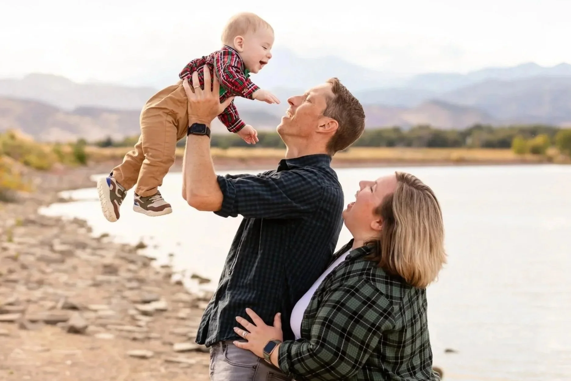 A family of three by a lake with mountains in the background, with a man lifting a smiling toddler boy dressed in plaid shirt, tan pants, and sneakers, while a woman, wearing a plaid shirt and watch, stands next to them looking up.