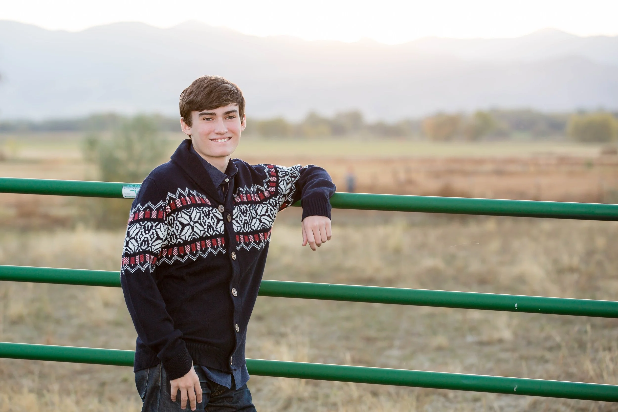 A rural high school senior portrait session featuring a young man in a knit cardigan leaning on a green cattle gate on a farm near McIntosh Lake in Longmont, Colorado, with views of Longs Peak. 
