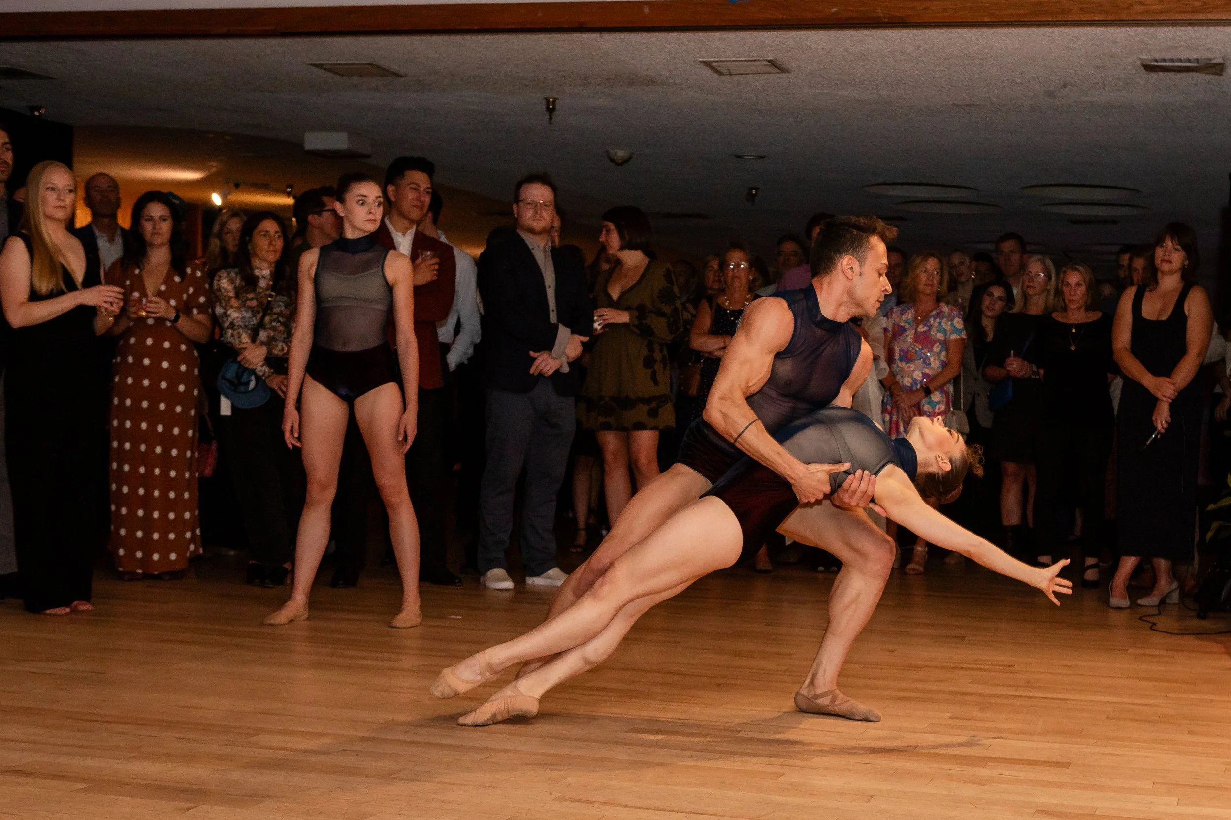 A group of people watching a ballet performance, with two dancers in the foreground engaged in a dance pose on a wooden floor.