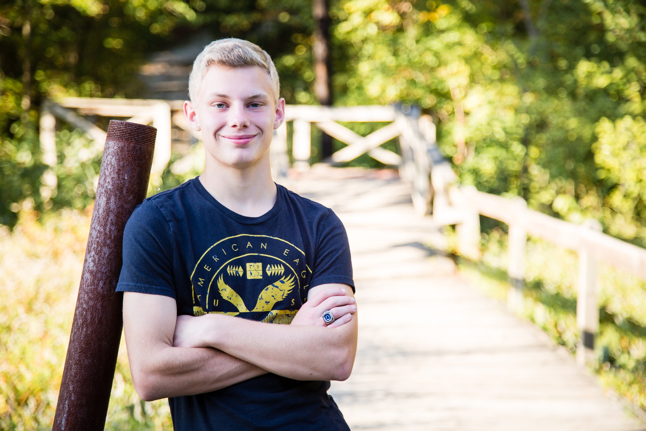 An outdoor high school senior portrait session featuring a young man leaning against a rustic structure with a wooden bridge and lush green foliage at Shakamak State Park in Jasonville, Indiana.