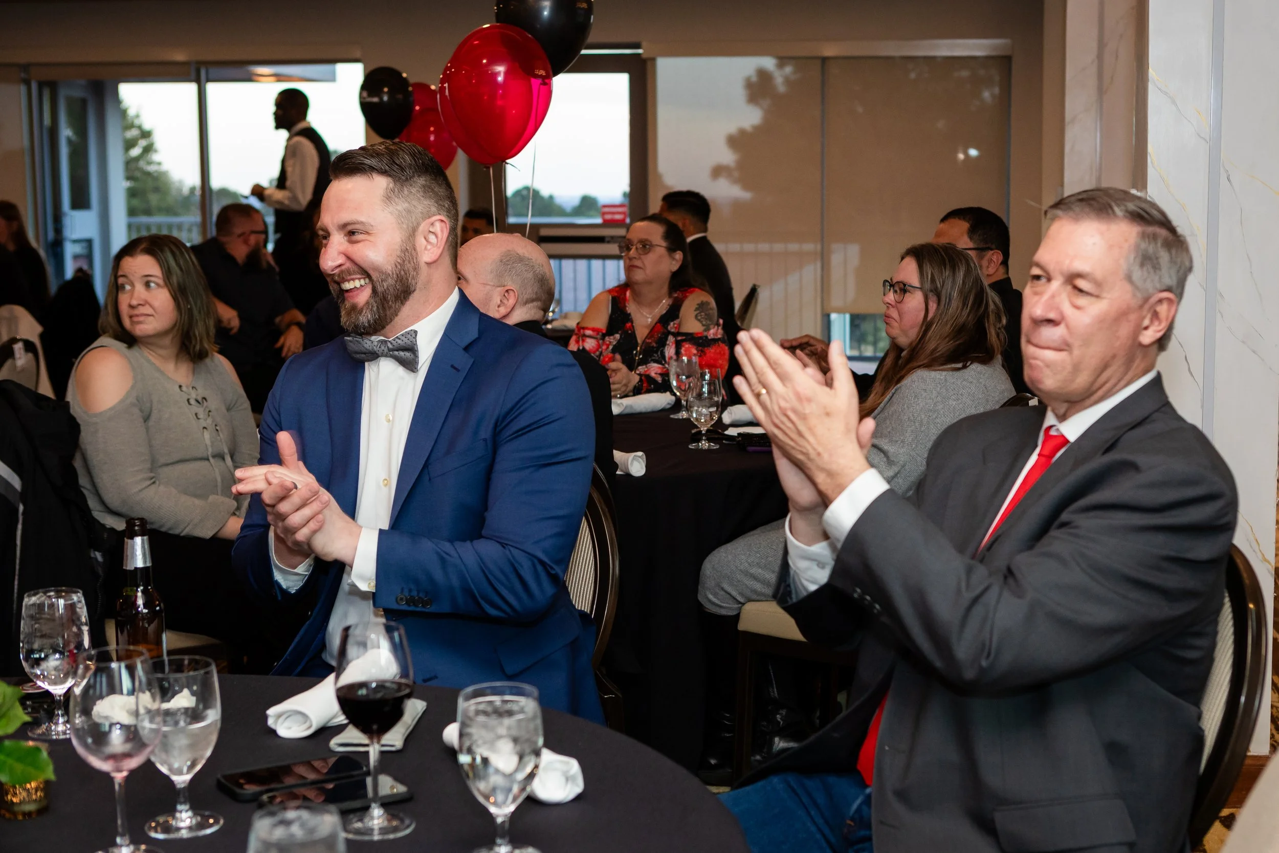 People sitting at a banquet table, some clapping and smiling, in a decorated room with balloons and large windows.