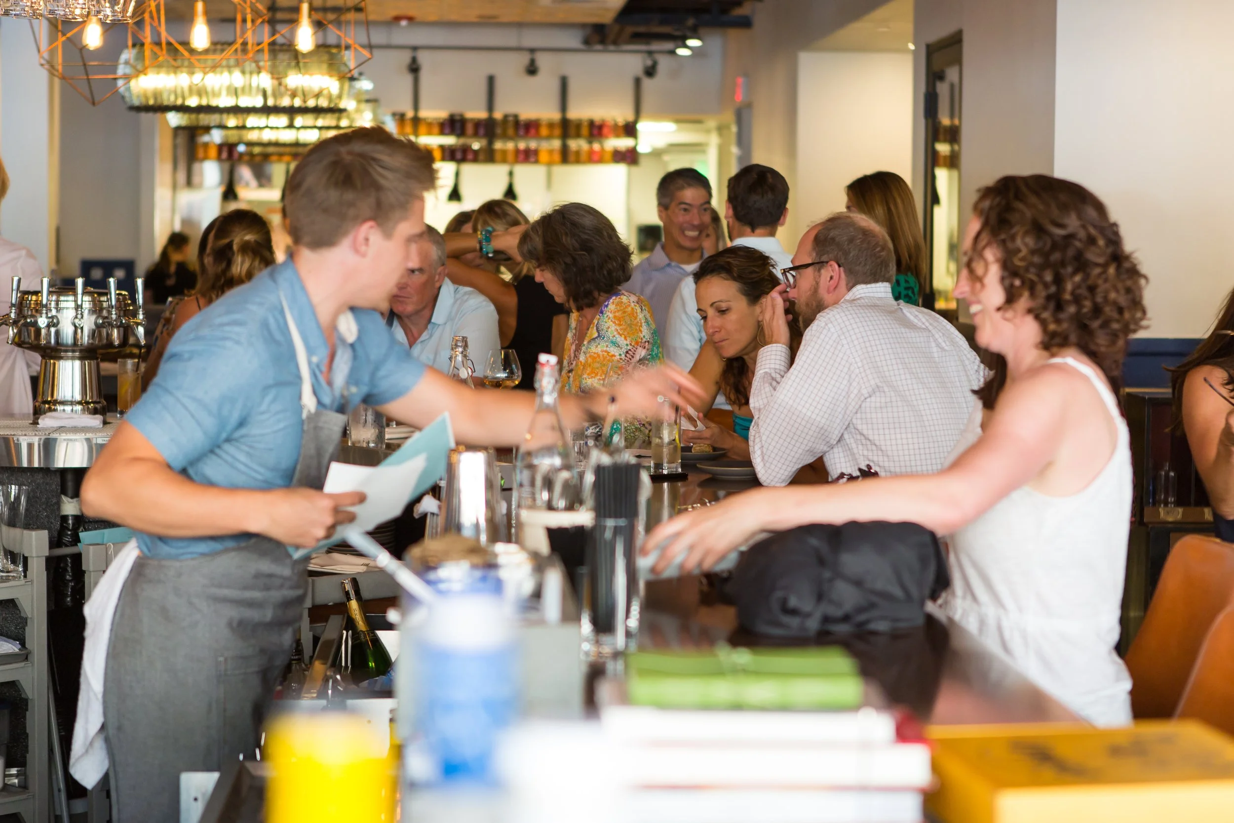 A busy restaurant scene with people seated at tables and a waiter in a blue shirt taking an order.