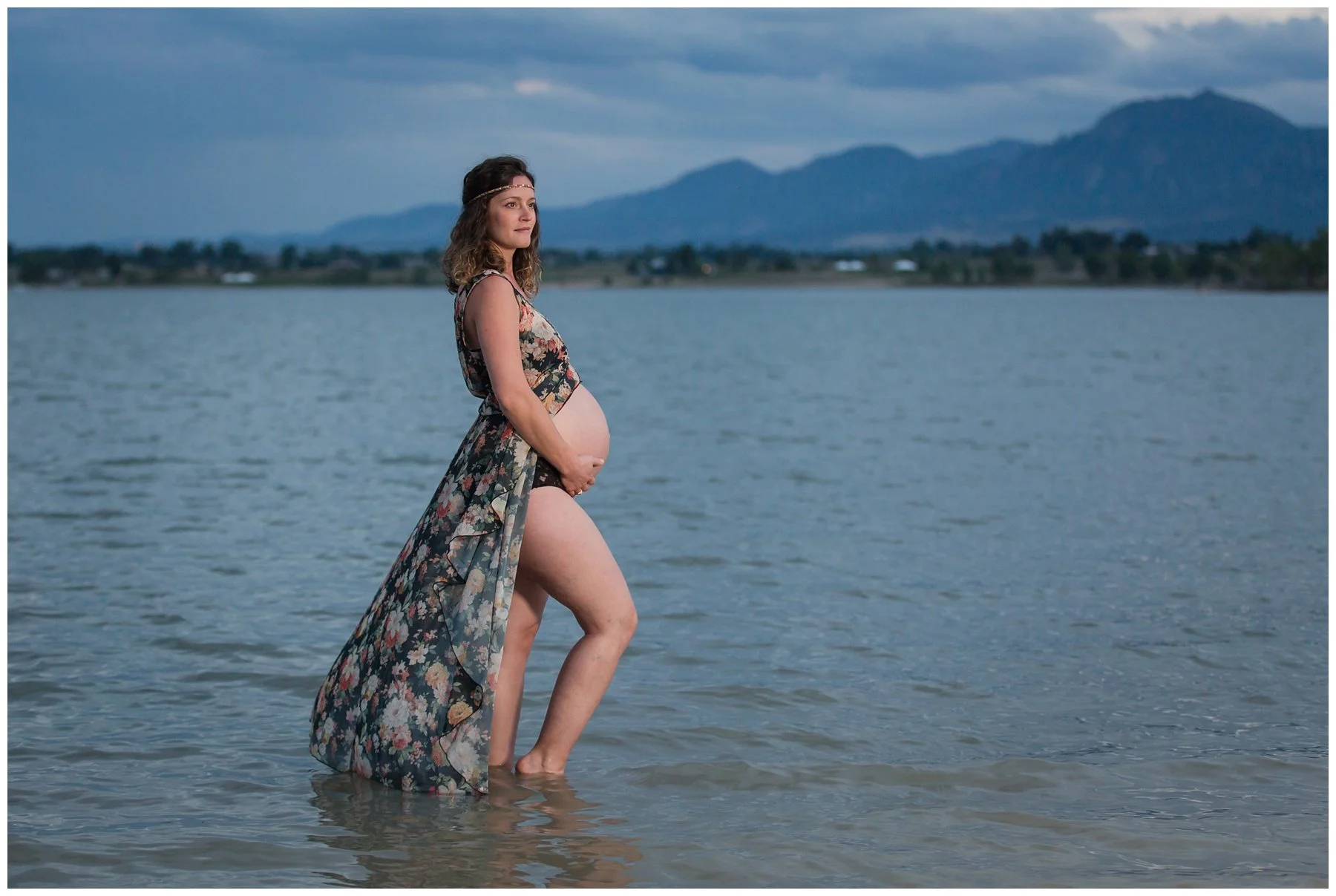 A pregnant woman wearing a floral dress and headband wades in a lake, with mountains and cloudy sky in the background.