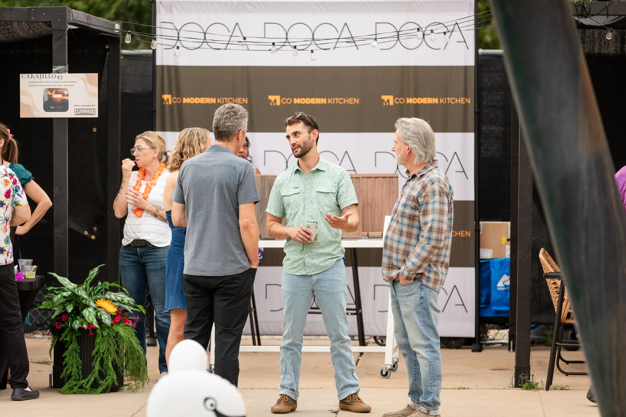 A group of people engaged in conversation at an outdoor event with a Food Doc Ada banner behind them. The setting includes a plant arrangement and hanging string lights.