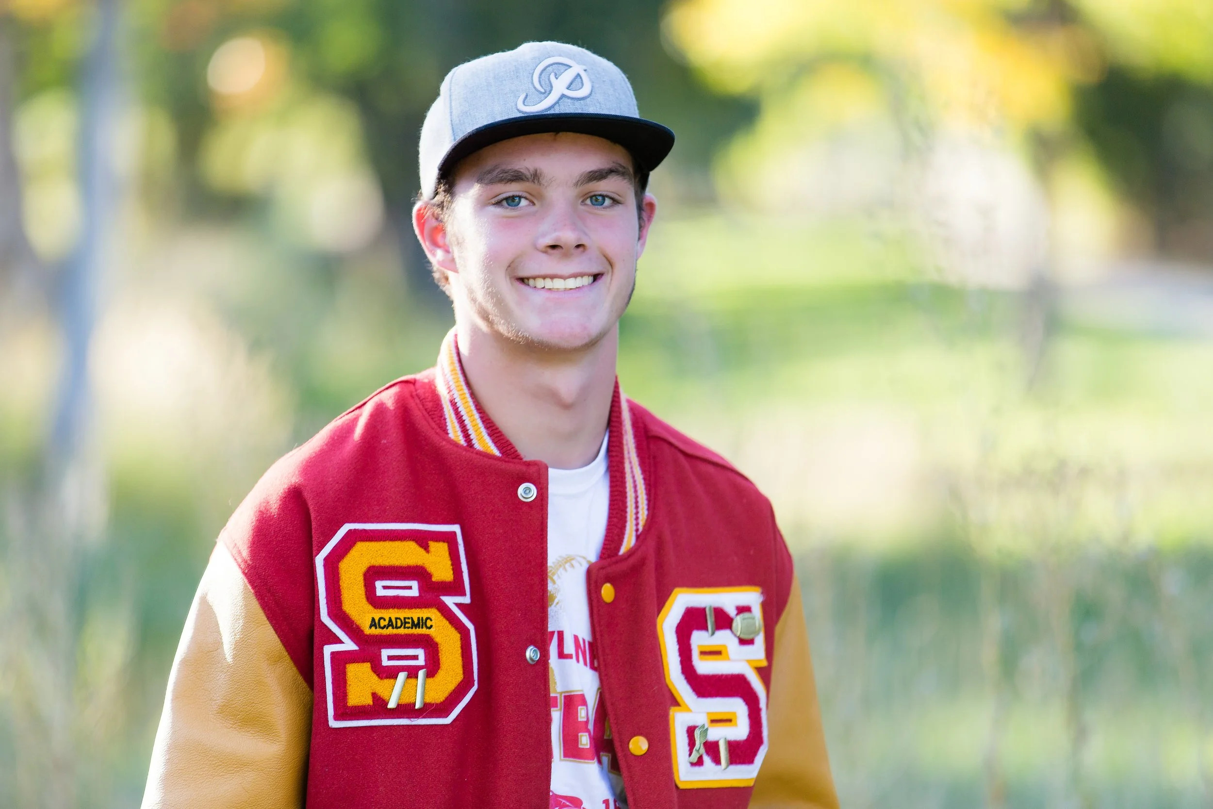 A young man with blue eyes and light brown hair wearing a gray cap and a red varsity jacket with yellow and red embroidered letters, standing outdoors with blurred trees and sunlight in the background.