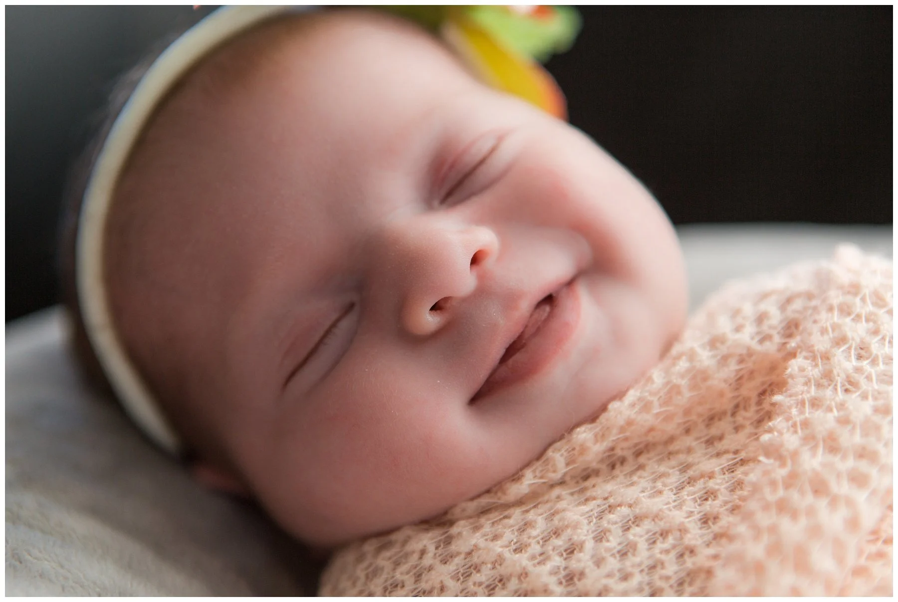 Close-up of a sleeping baby with smooth skin, wearing a headband and wrapped in a soft, peach-colored blanket.