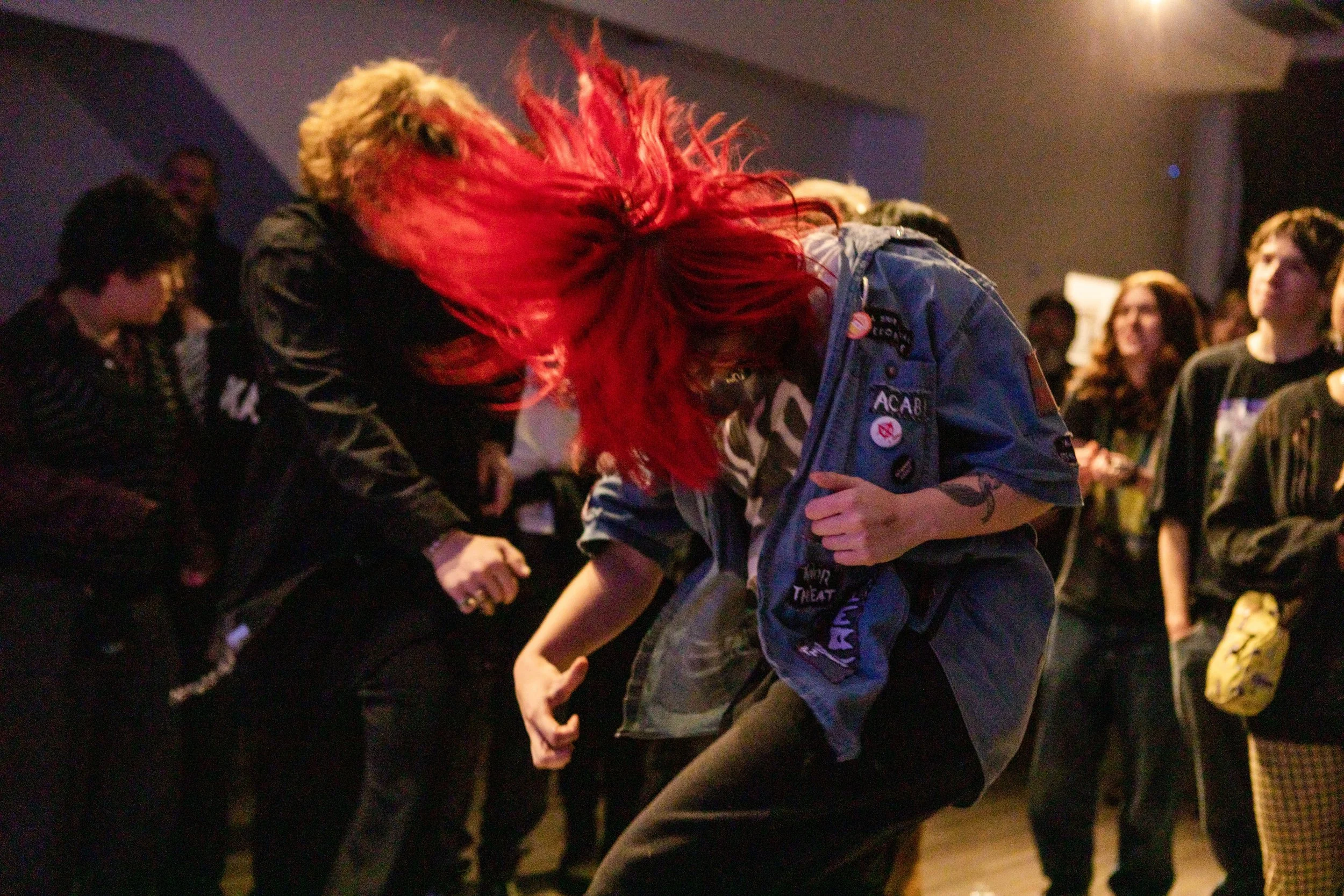 A group of people dancing together at a dance party or social event, with a woman with vibrant red hair and a denim jacket in the center, surrounded by others enjoying the moment.