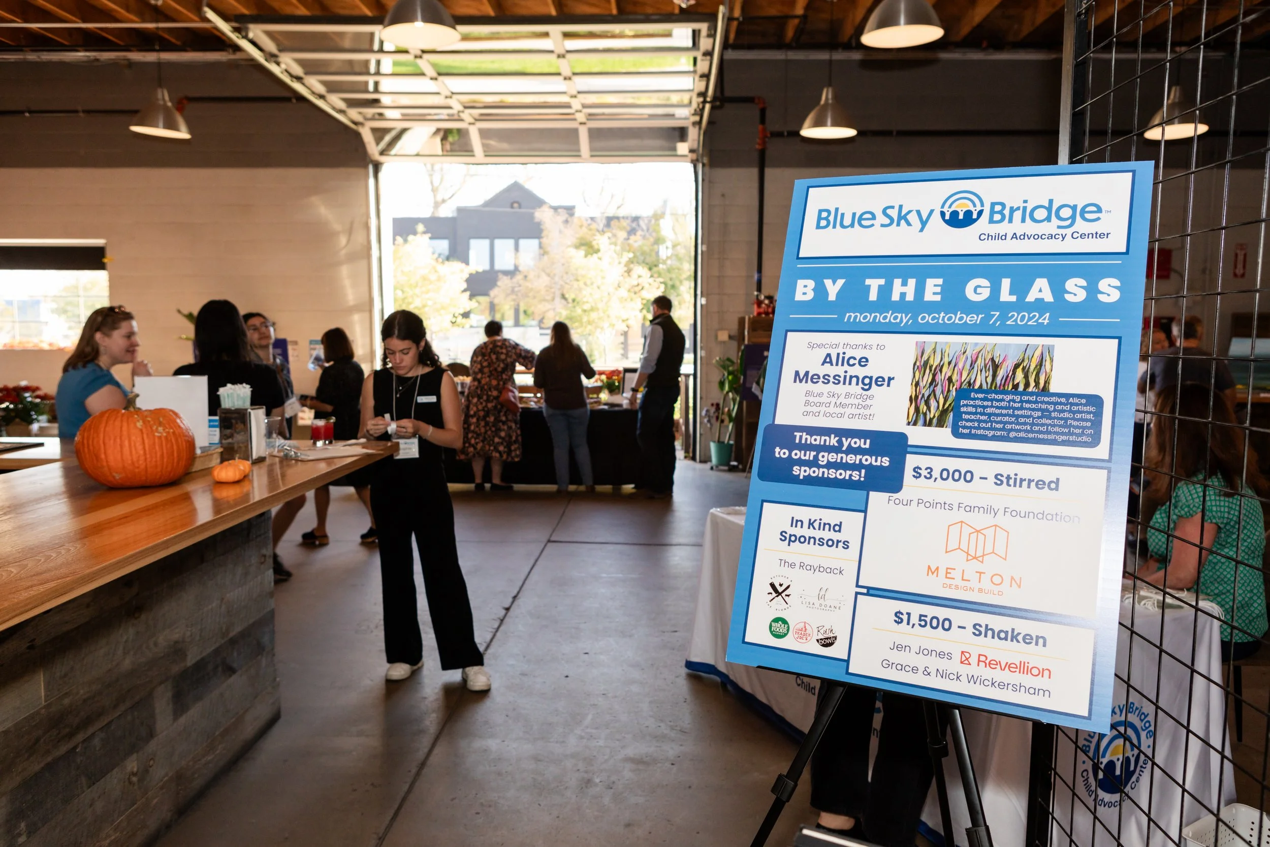 People at a community event with a sign for Blue Sky Bridge Child Advocacy Center, featuring sponsors and event details, inside a building with high ceilings and natural light.