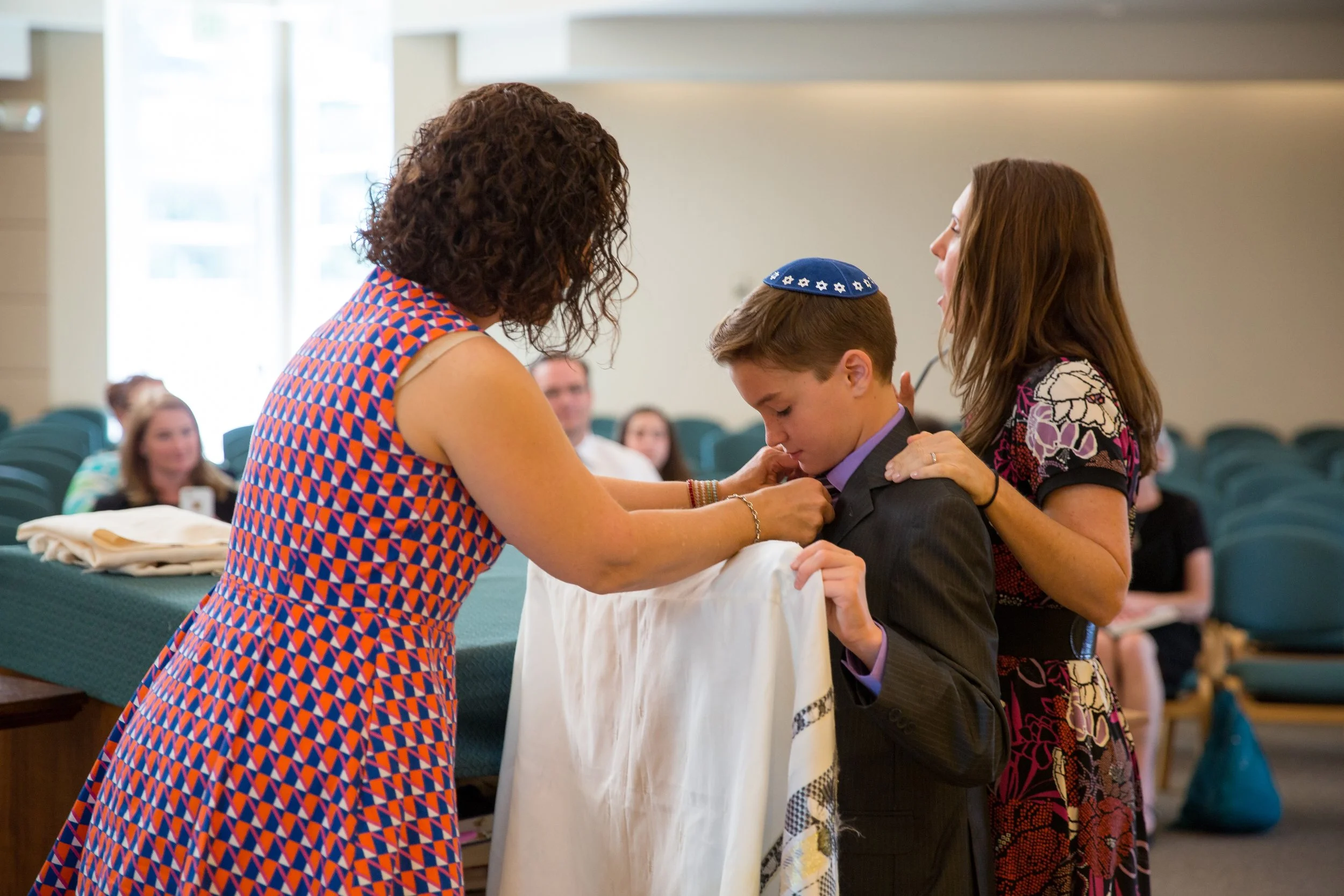 An intimate documentary style photograph capturing a family helping a young boy adjust his tallit during a Bar Mitzvah ceremony rehearsal, by Lisa Doane Photography.