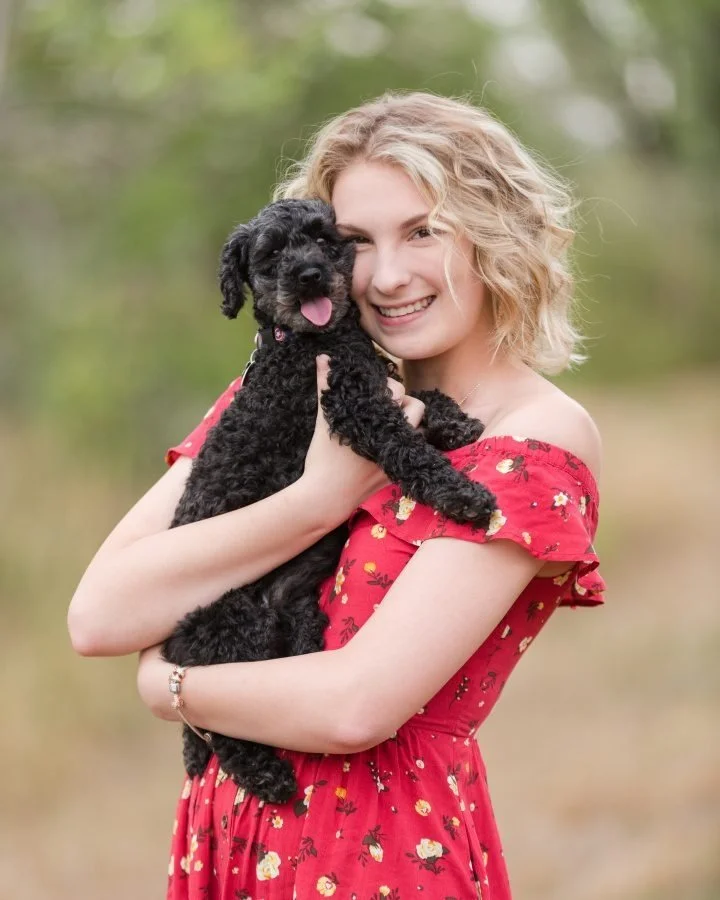 A young woman with blonde curly hair in a red floral dress holding a black puppy outdoors with green trees in the background.