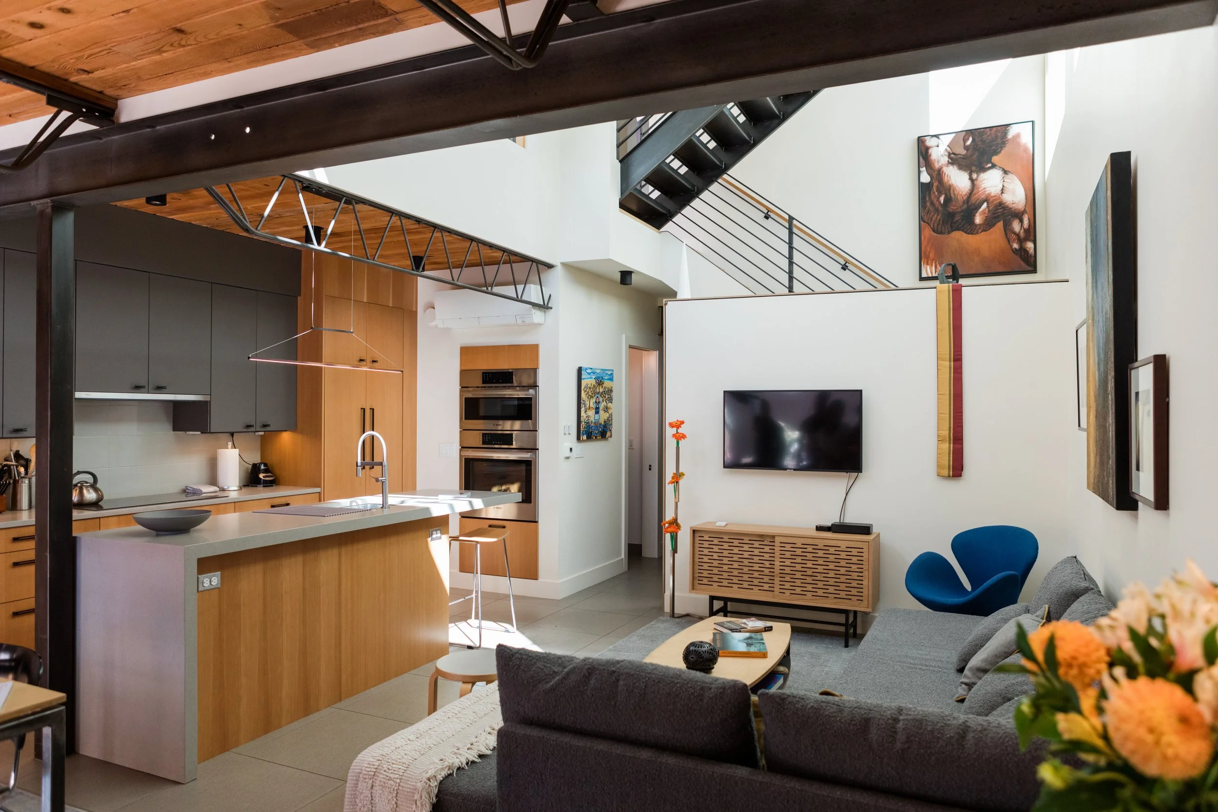 Modern open-plan kitchen and living room with vaulted ceiling, black staircase, abstract art, and colorful decor.