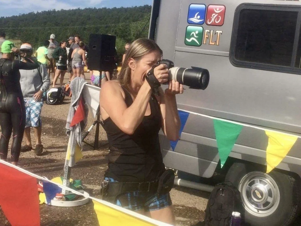 Woman takes a photograph with a professional camera at an outdoor event, with other people in the background and a van decorated with colorful flags.