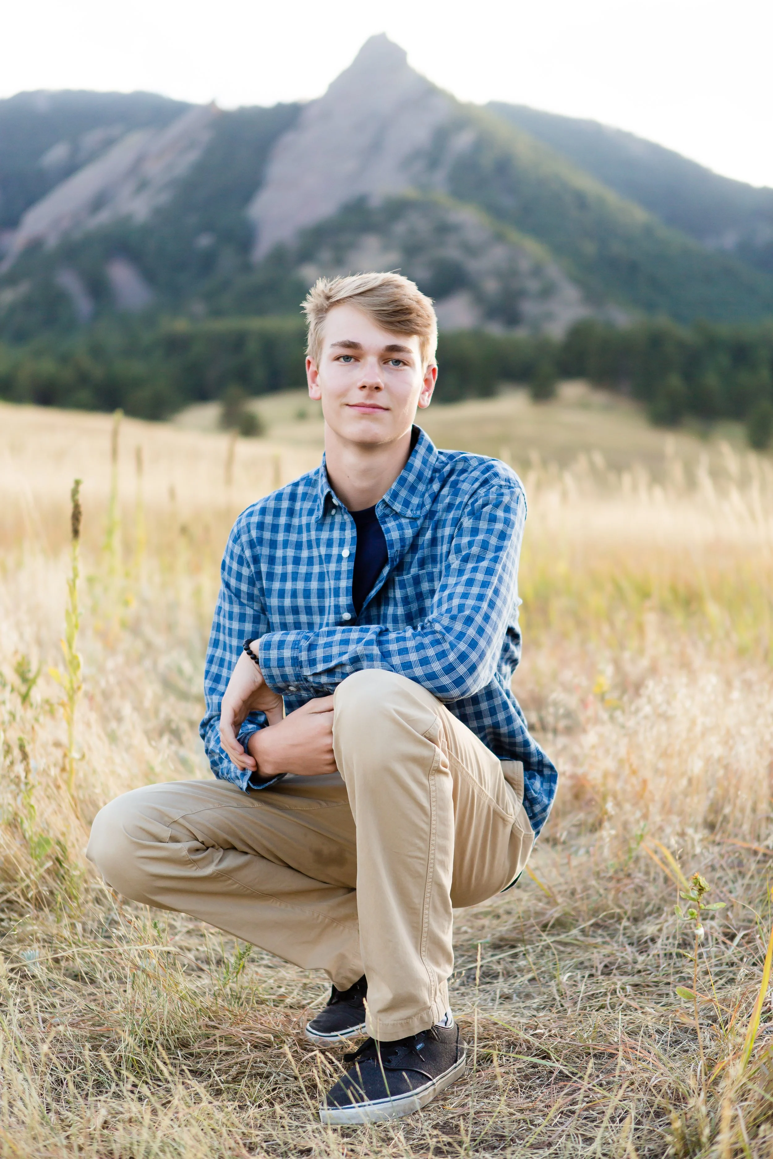 Young man with blond hair wearing a blue plaid shirt and beige pants, crouching in a field of tall grass with mountains in the background.