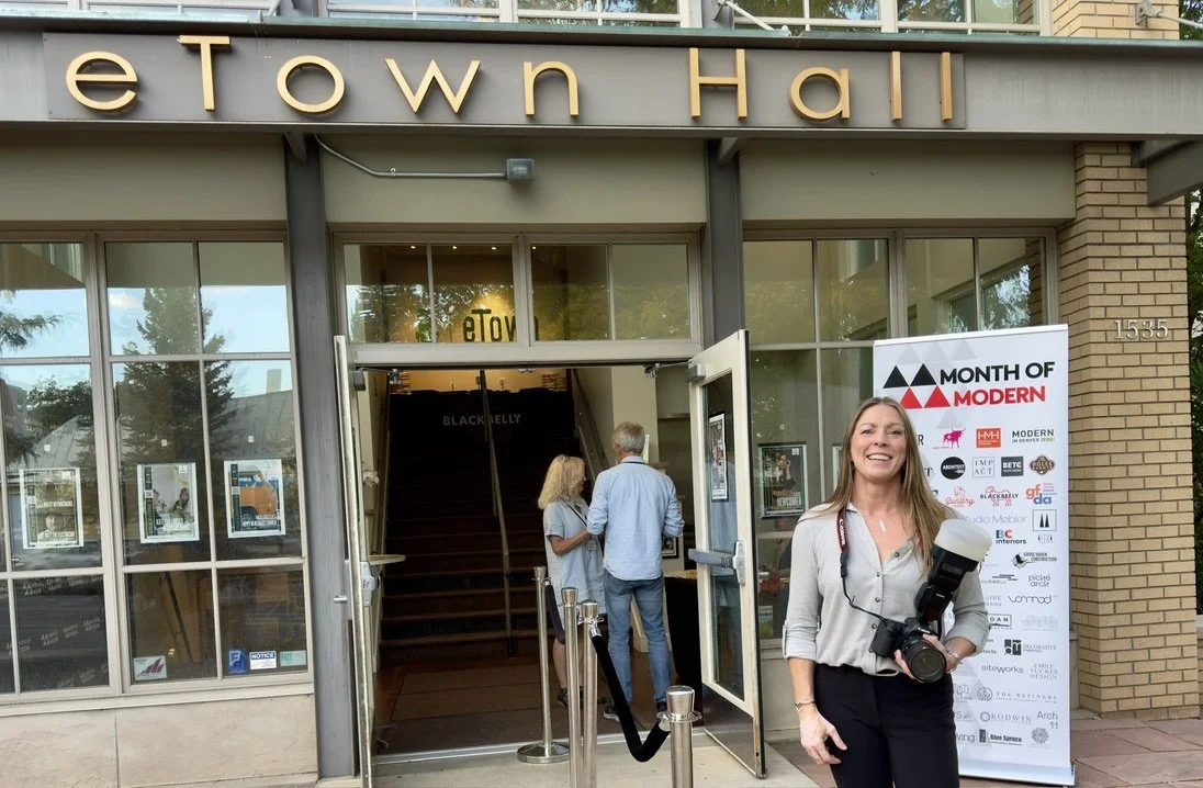 A woman standing outside Town Hall with a camera and a professional flash, smiling at the camera. Behind her, there are people entering the building and a promotional banner on the right side that reads 'Month of Modern.' The building has large glass windows and a staircase visible through the open doors.