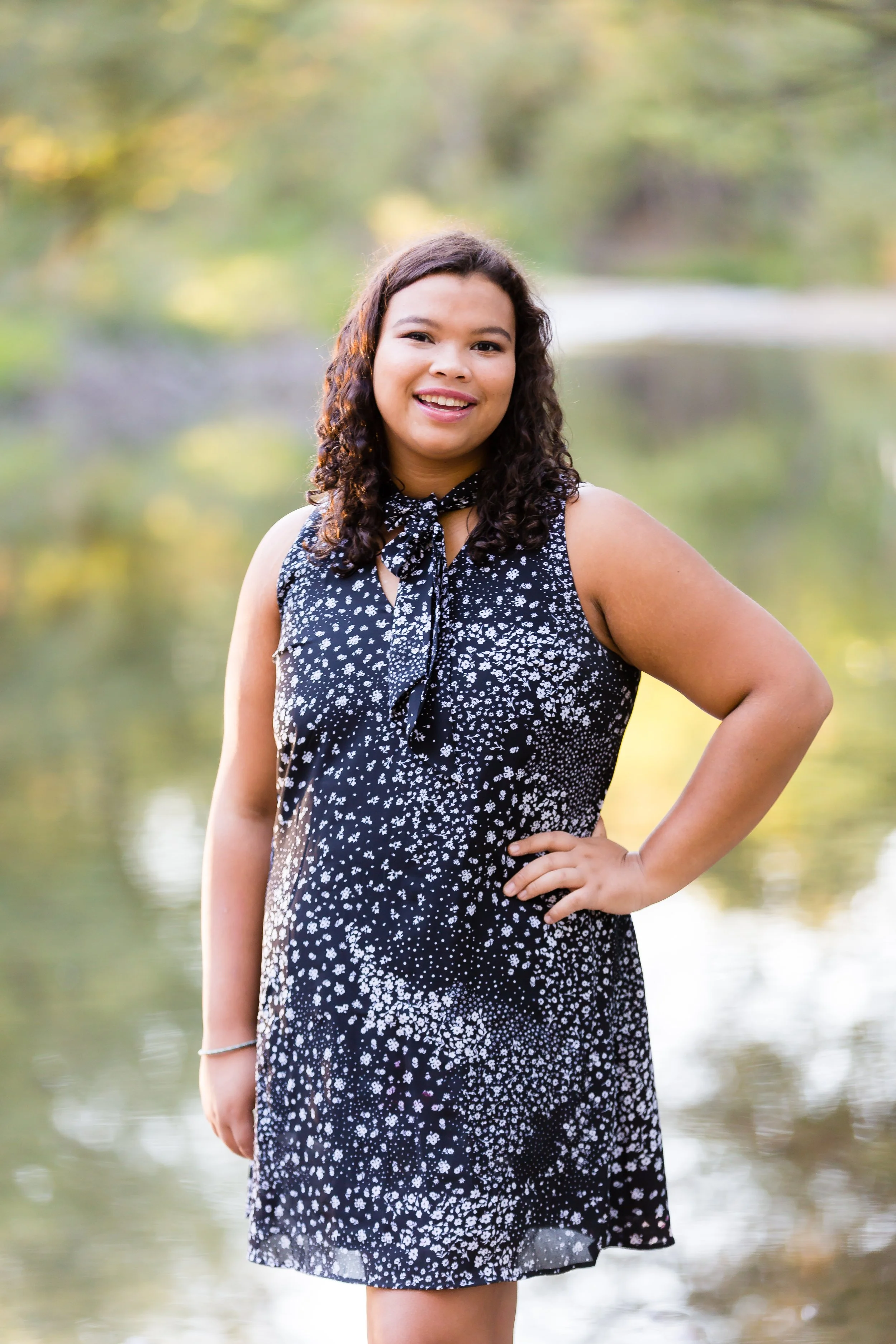 A young woman with dark, curly hair stands outdoors next to a body of water, smiling at the camera during daytime.