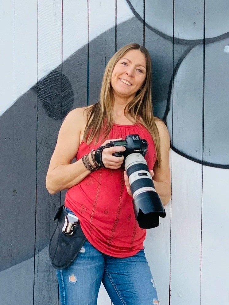 Professional portrait of Lisa Doane, owner of Lisa Doane Photography, holding a camera with a telephoto lens while leaning against an urban mural.