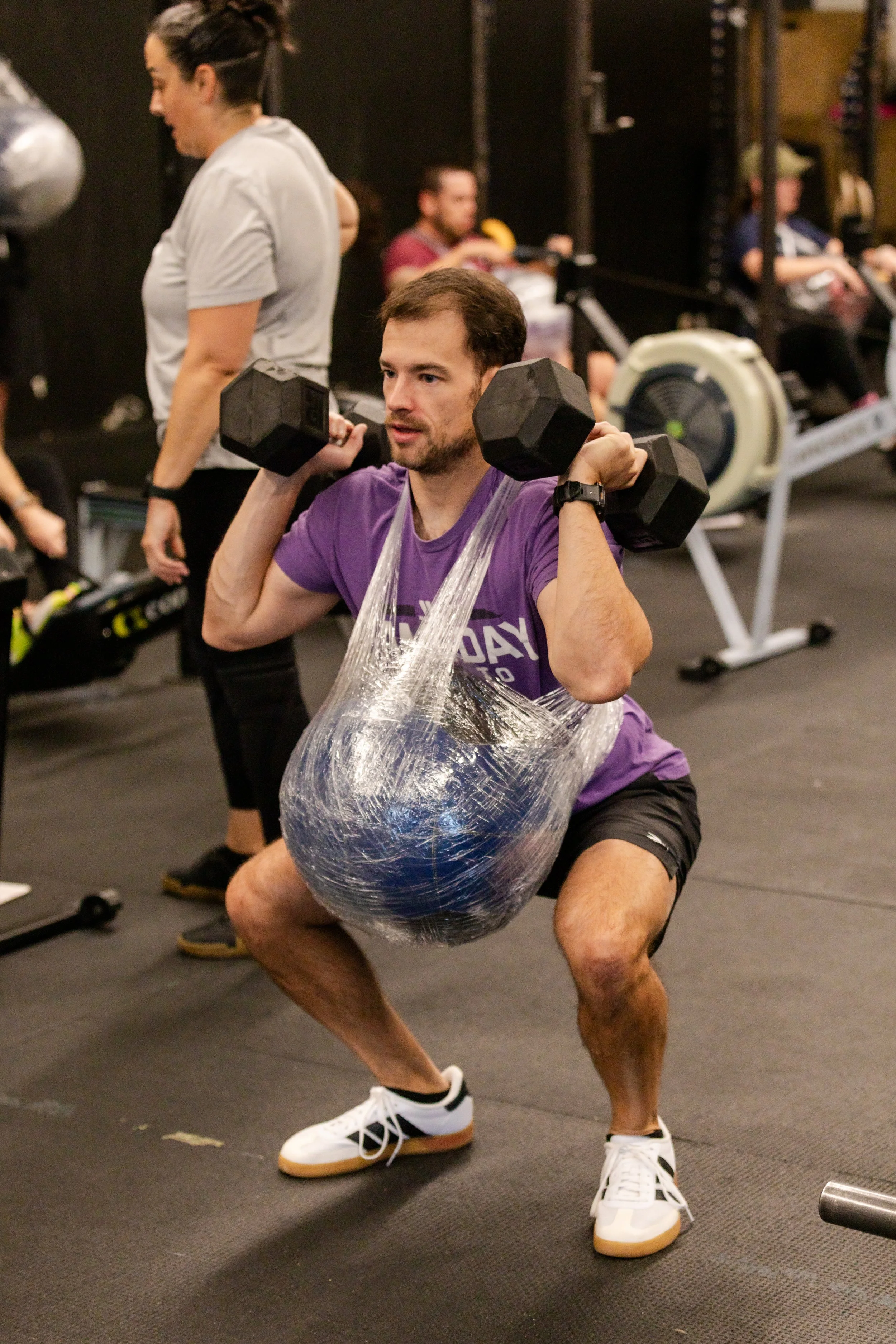 A man performing a squat exercise with two dumbbells on his shoulders, wrapped in plastic wrap, in a gym. He is wearing a purple shirt, black shorts, and white sneakers. In the background, other people are working out and exercising on equipment.