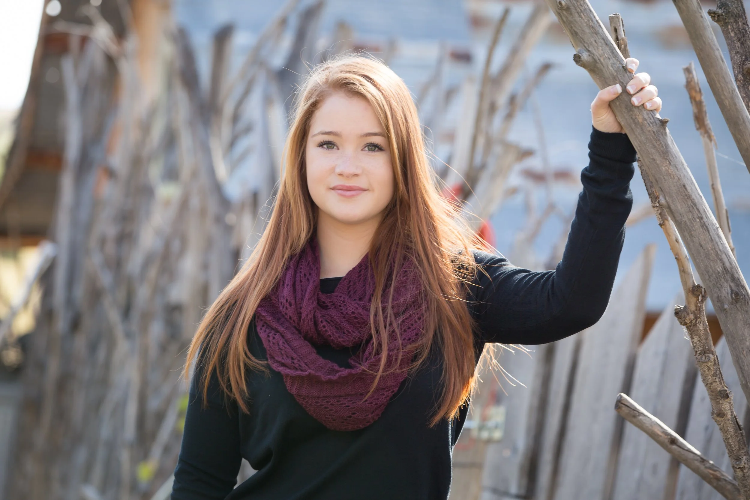 An outdoor high school senior portrait session featuring a young woman posing next to a homemade rustic branch fence on location at Lone Hawk Farm in Longmont, Colorado, by Lisa Doane Photography.