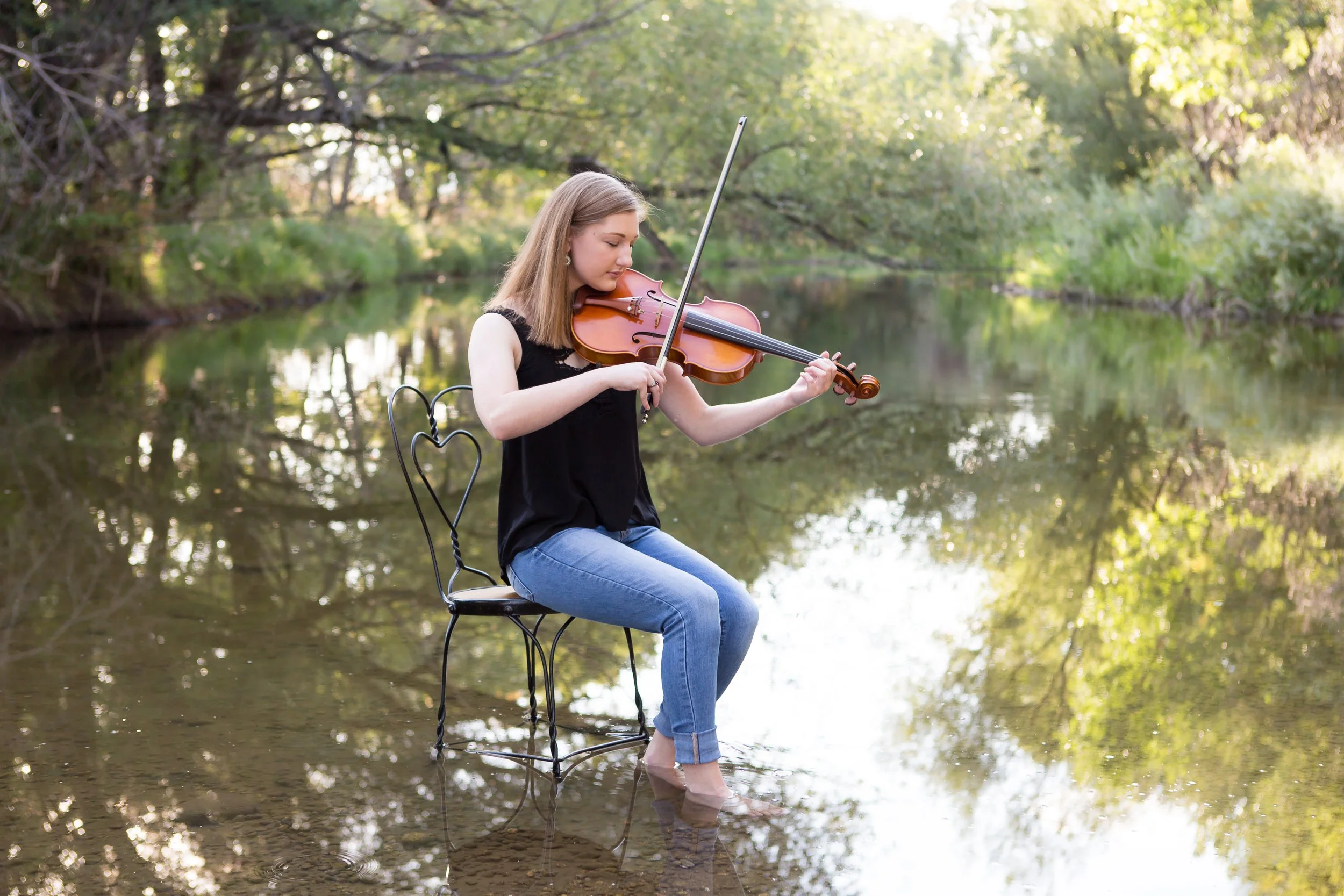 A young woman with blonde hair, wearing a black sleeveless top and blue jeans, sitting on a metal chair with a heart-shaped backrest in shallow water by a river or pond, playing the violin with a forested background.